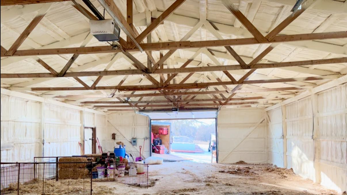 Interior of a barn with wooden trusses, white walls, tools, and two people near the far open doorway.