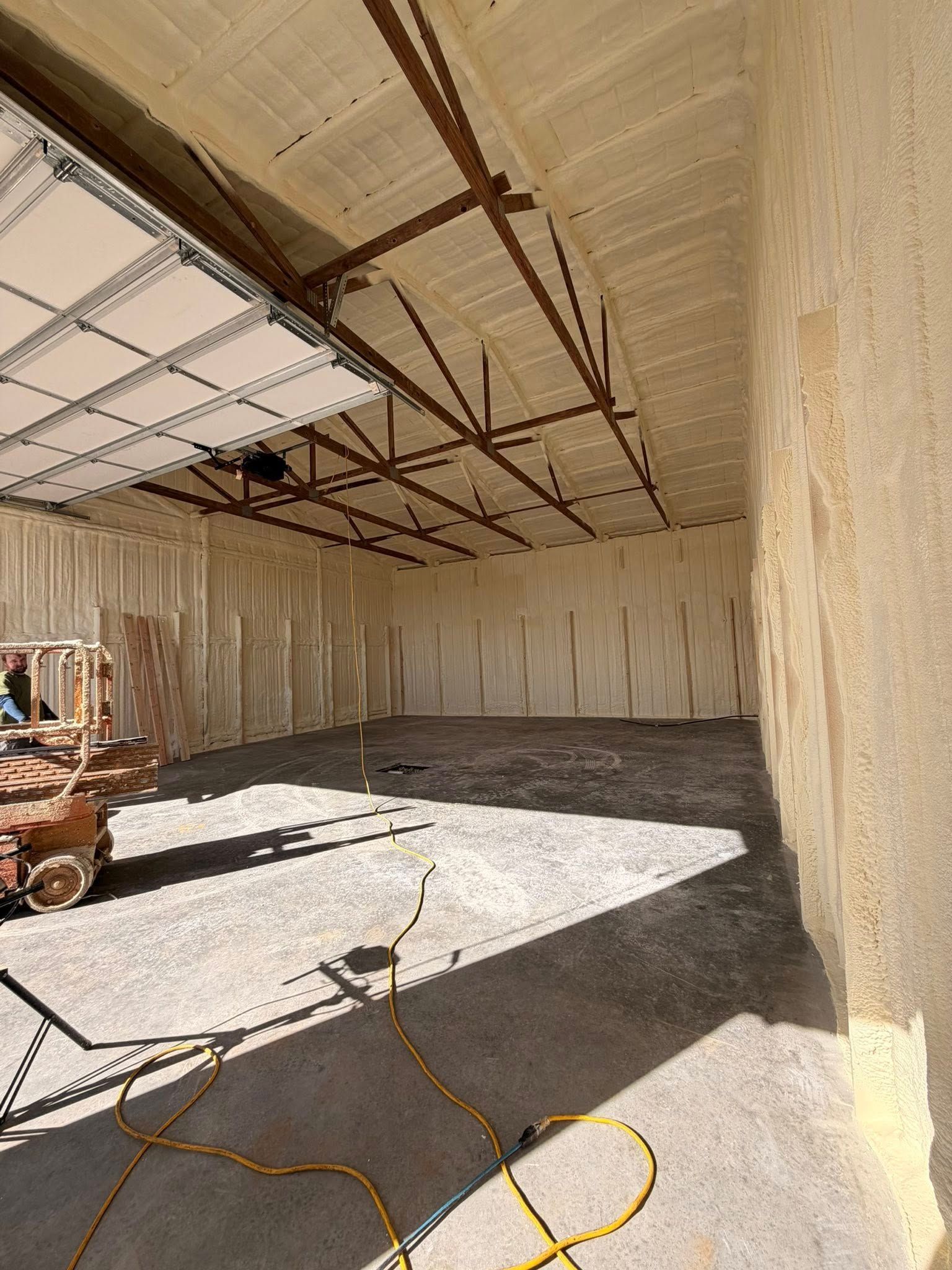 Empty concrete room under construction with high insulated walls and ceiling trusses, sunlight on floor, and cords scattered.