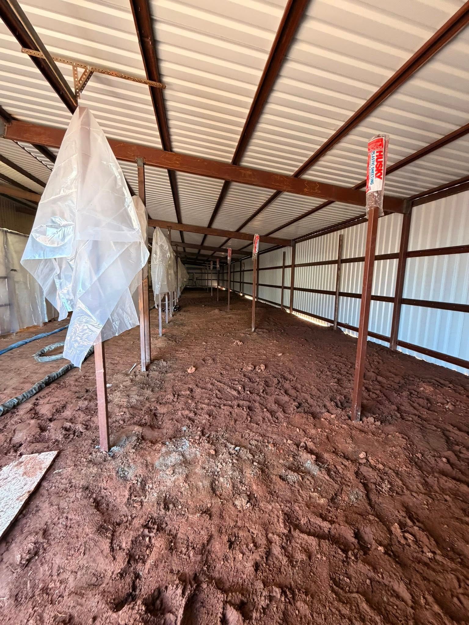 Empty barn interior with red dirt floor, metal roof, and a plastic-covered post on the left