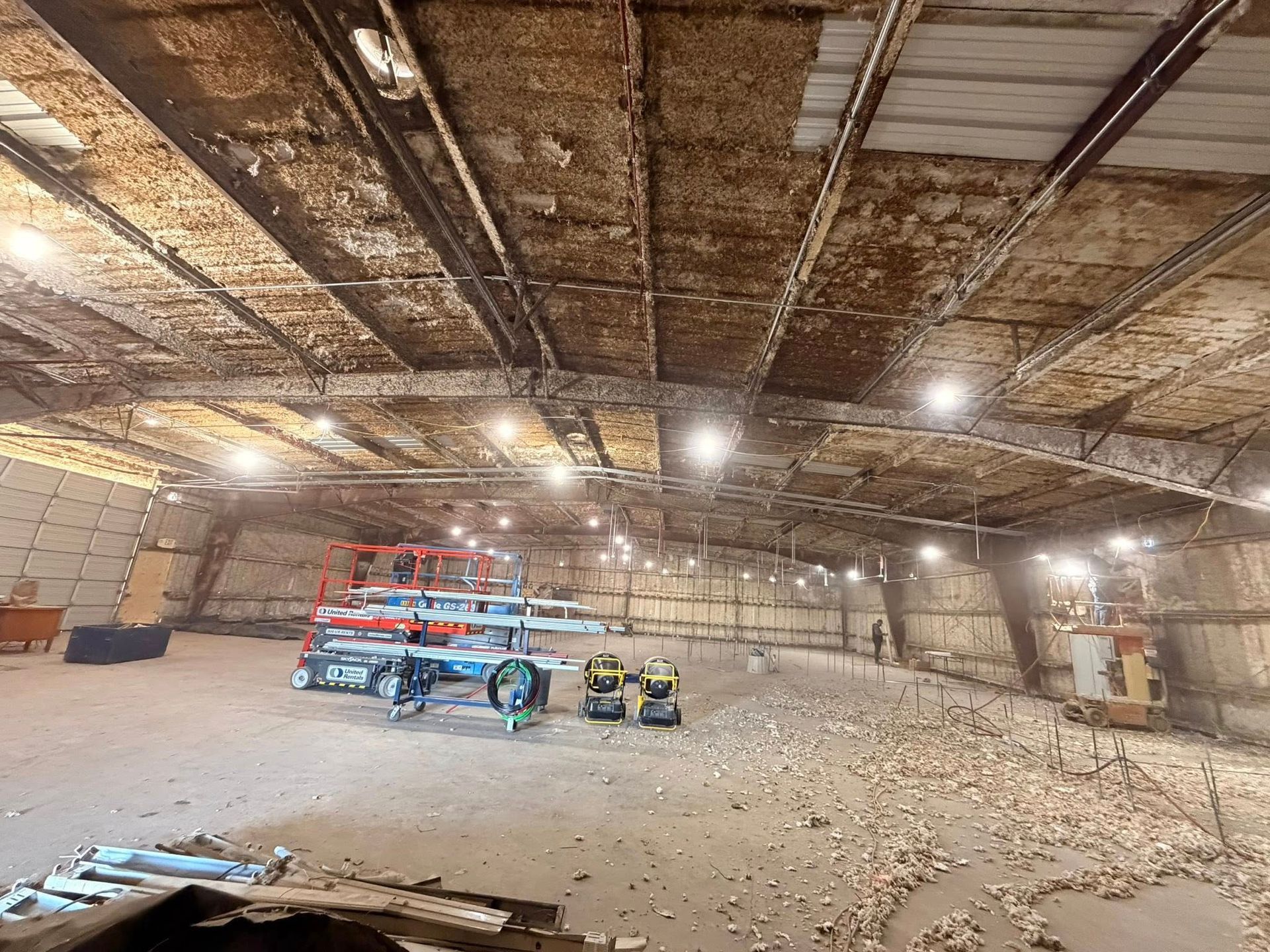 Large empty warehouse with exposed ceiling, scattered debris, and a few vehicles and equipment inside.