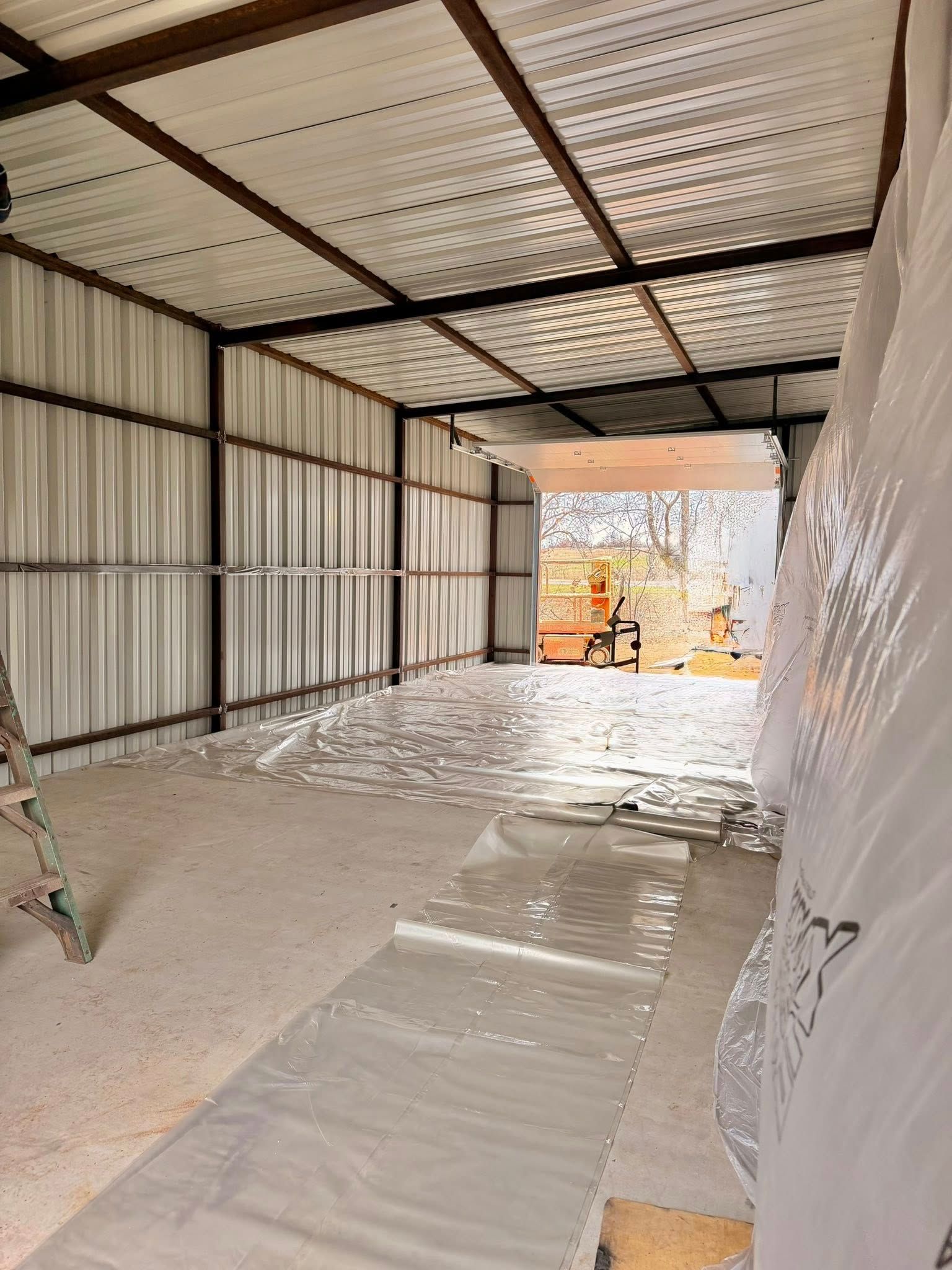 Empty corrugated-metal shed with a dirt floor and plastic sheeting covering the ground, viewed toward an open doorway.