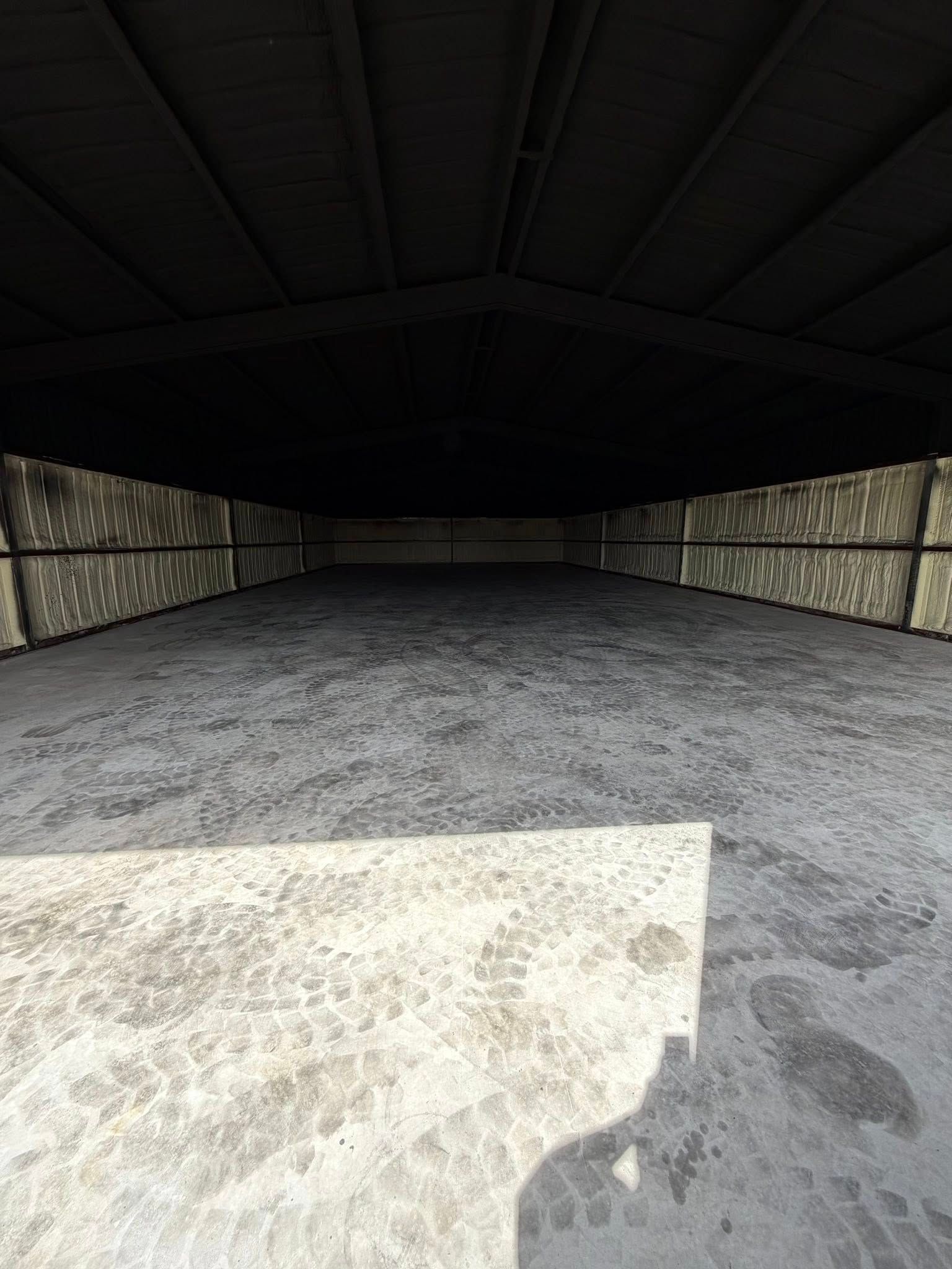 Empty indoor driving range with a dark ceiling and gravel floor, lit by daylight at the open sides
