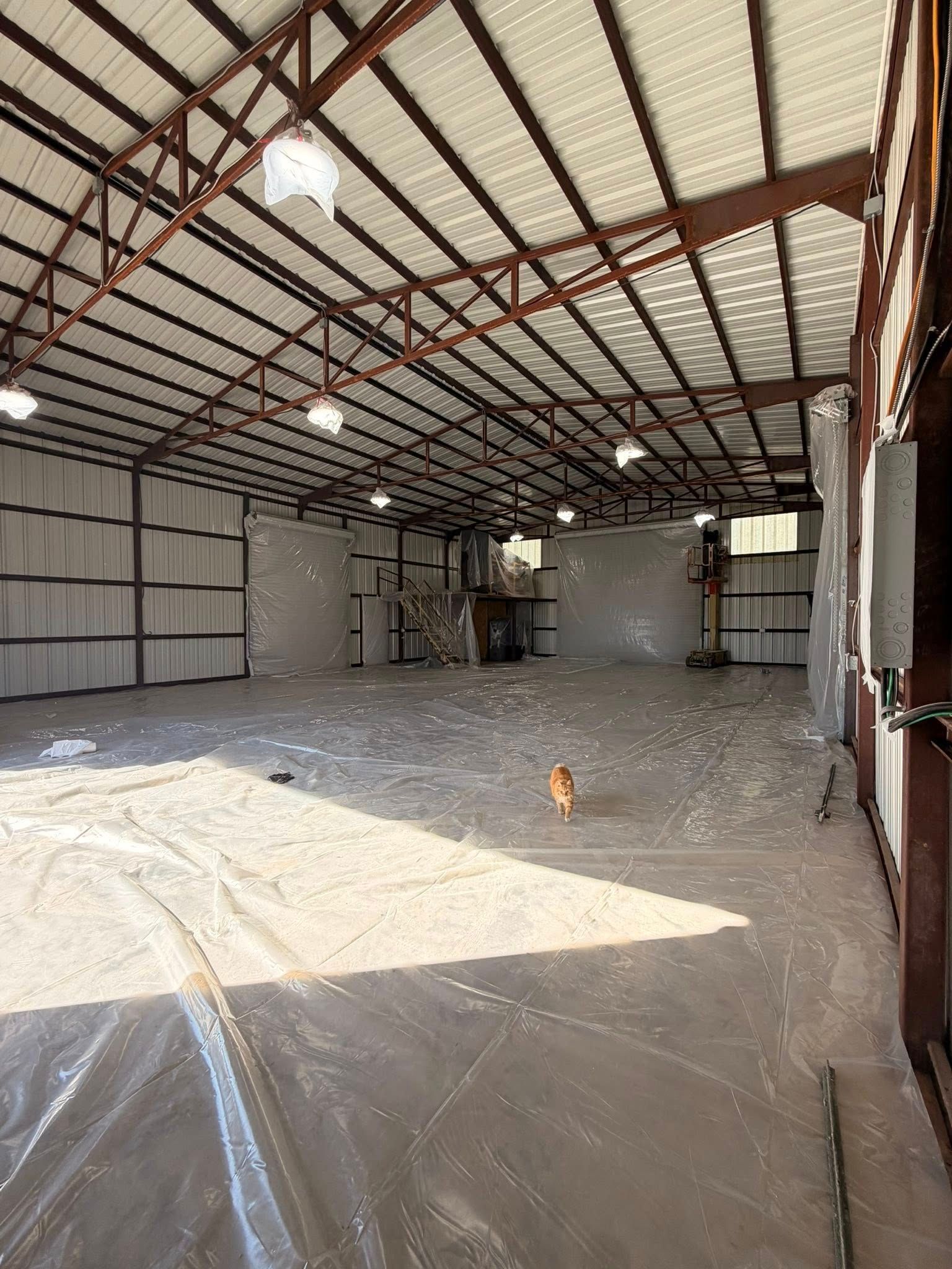 Empty metal warehouse with a high roof, concrete floor, and plastic sheeting covering part of the ground.