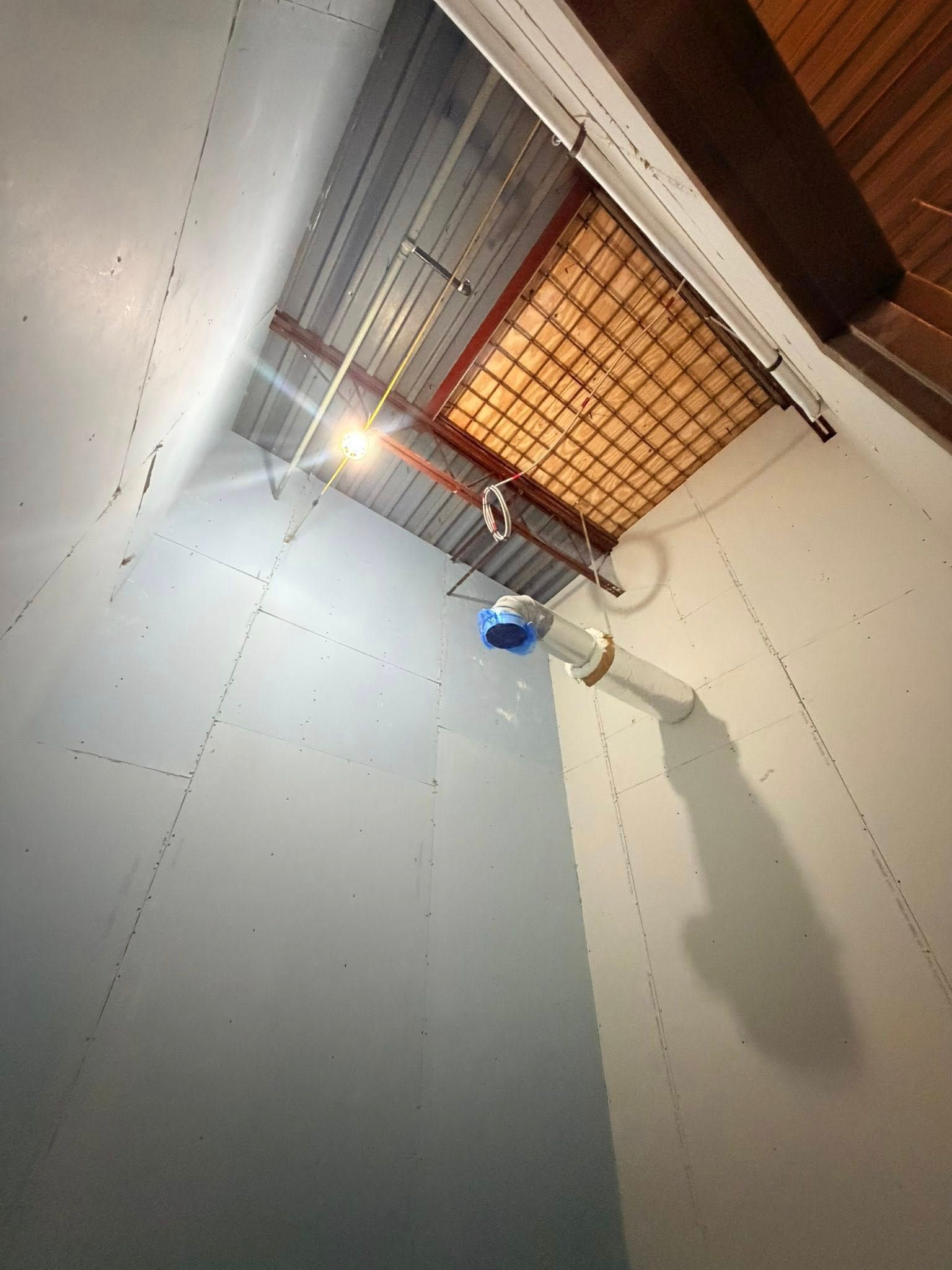 Ceiling vent with orange grille, exposed pipes, and a blue device hanging below in a white utility space