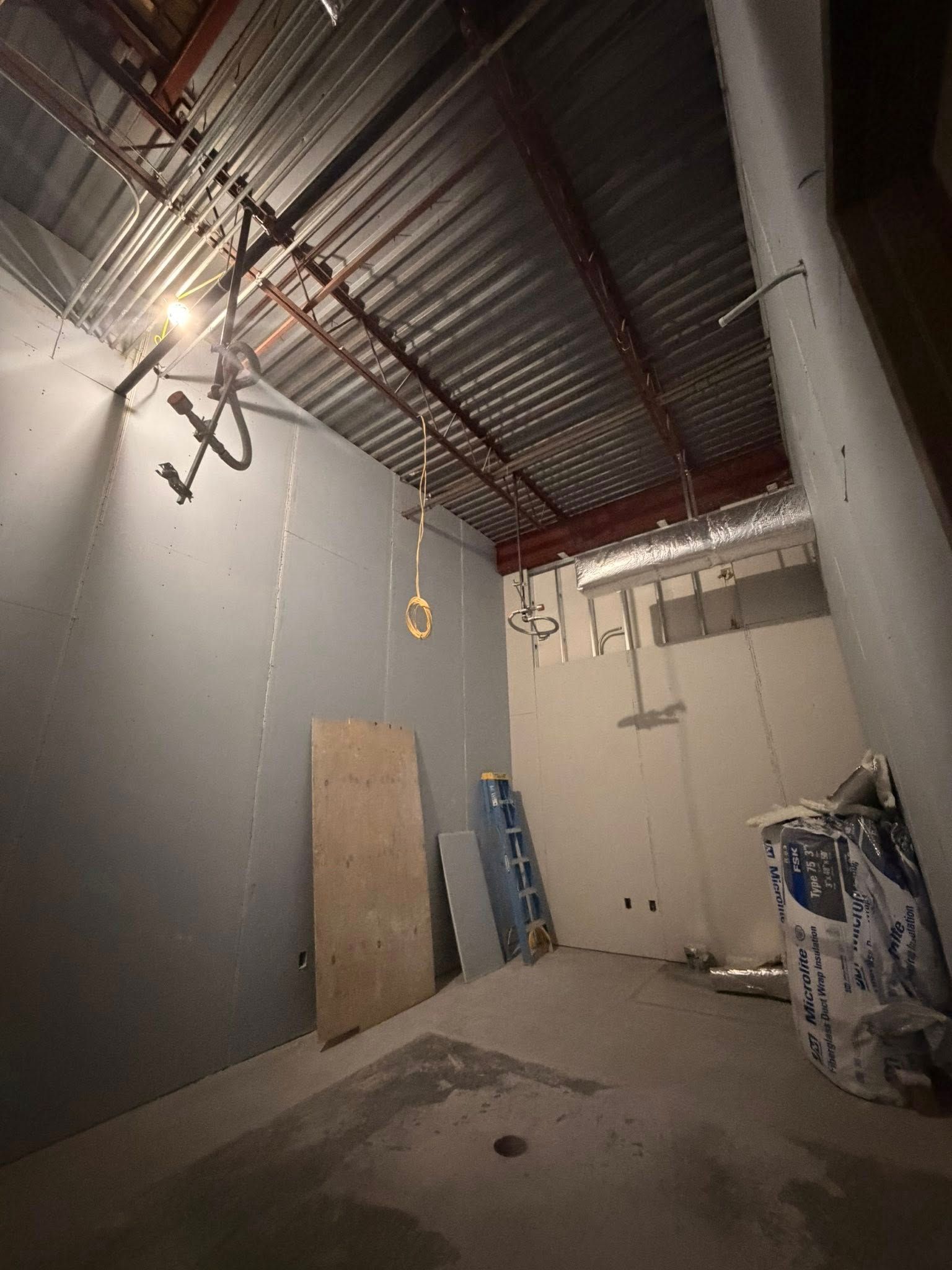 Empty industrial room with exposed ceiling ducts, gray walls, and construction materials leaning against the walls