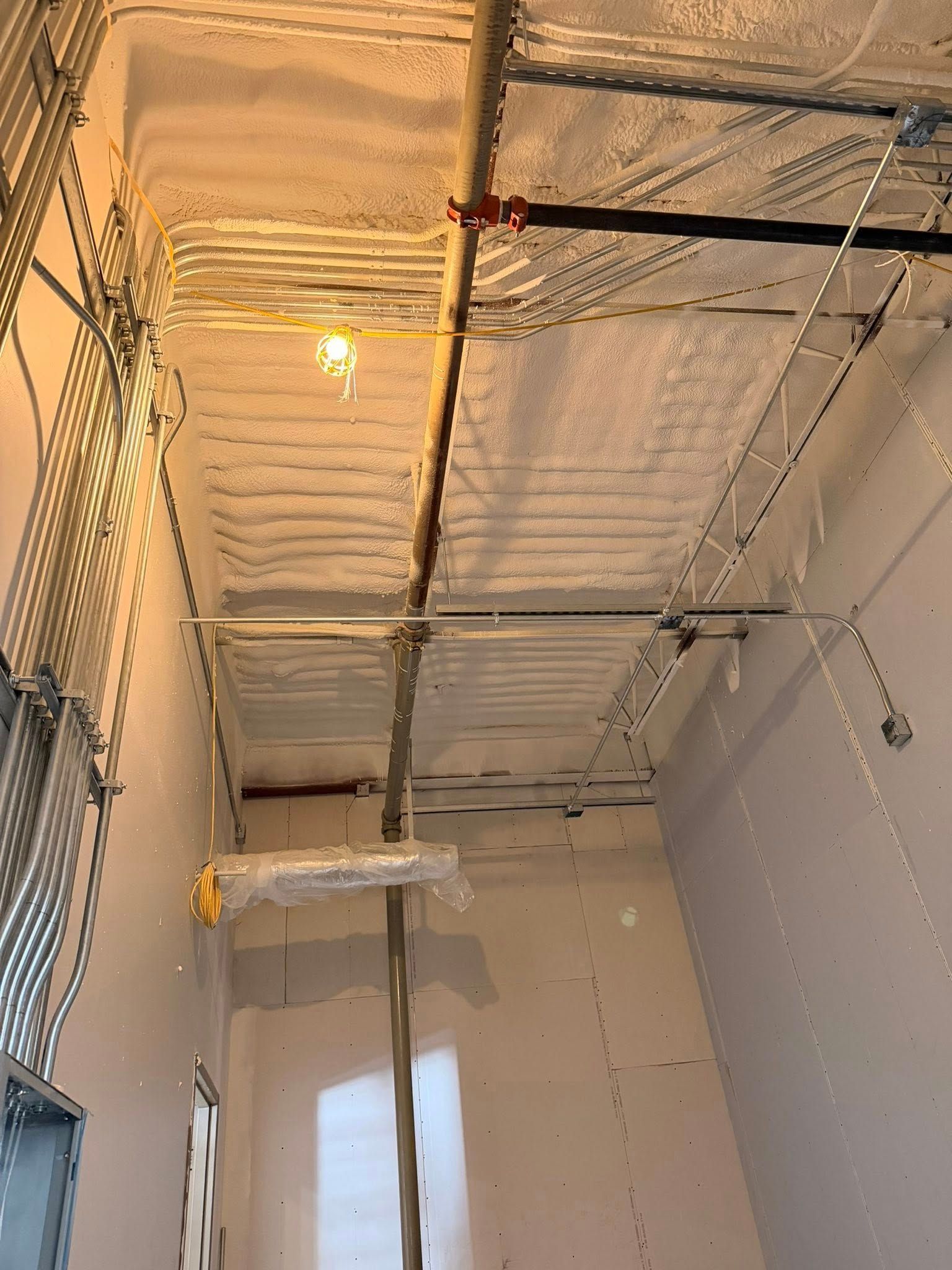 Ceiling with exposed pipes and insulation above a small white room, lit by a single bulb.
