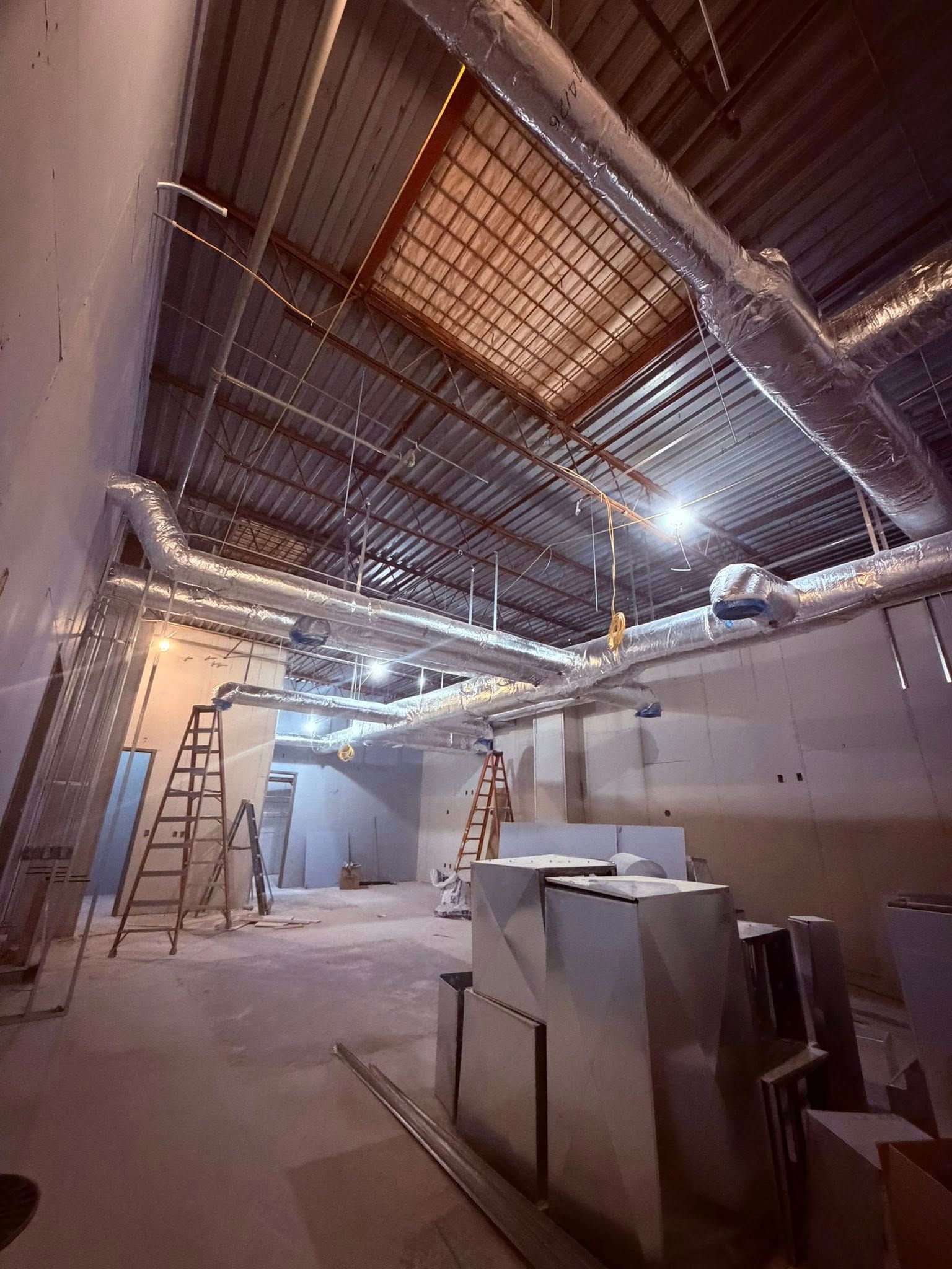 Industrial interior under construction with exposed ceiling grids, ladders, ductwork, and worktables.
