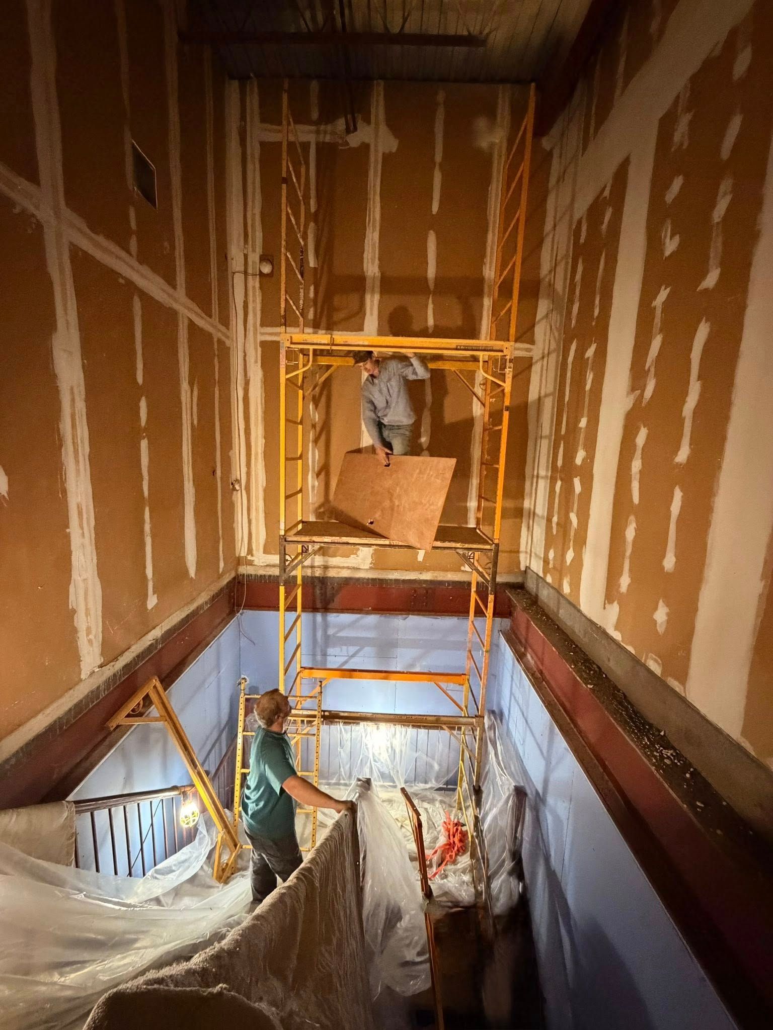 Scaffold in unfinished interior stairwell with workers below and drywall walls.