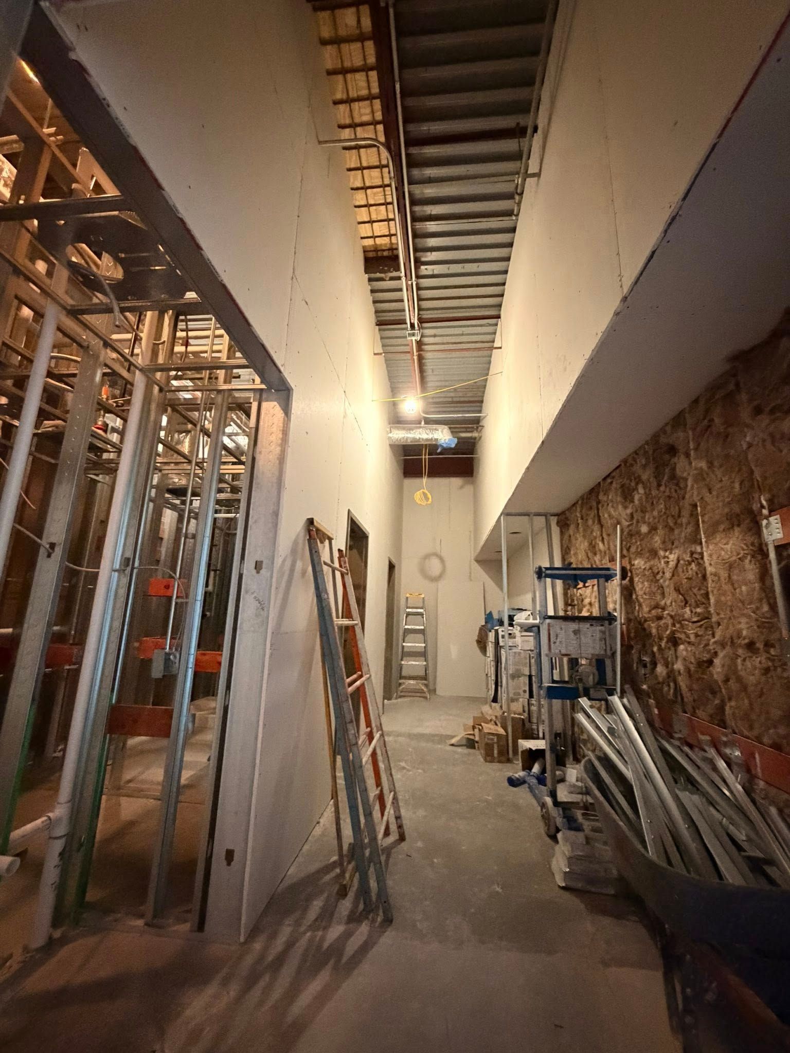 Narrow construction corridor with exposed framing, ladders, and stacked materials under ceiling work lights