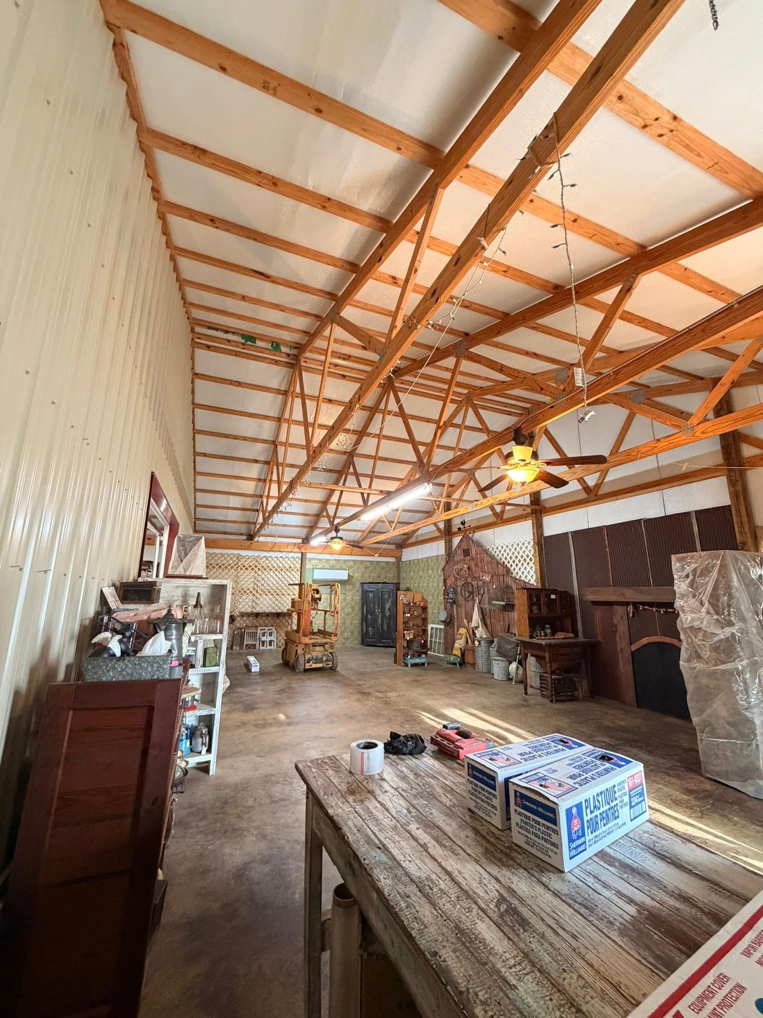 Wide indoor workshop with exposed wooden trusses, long tables, shelves, and hanging lights