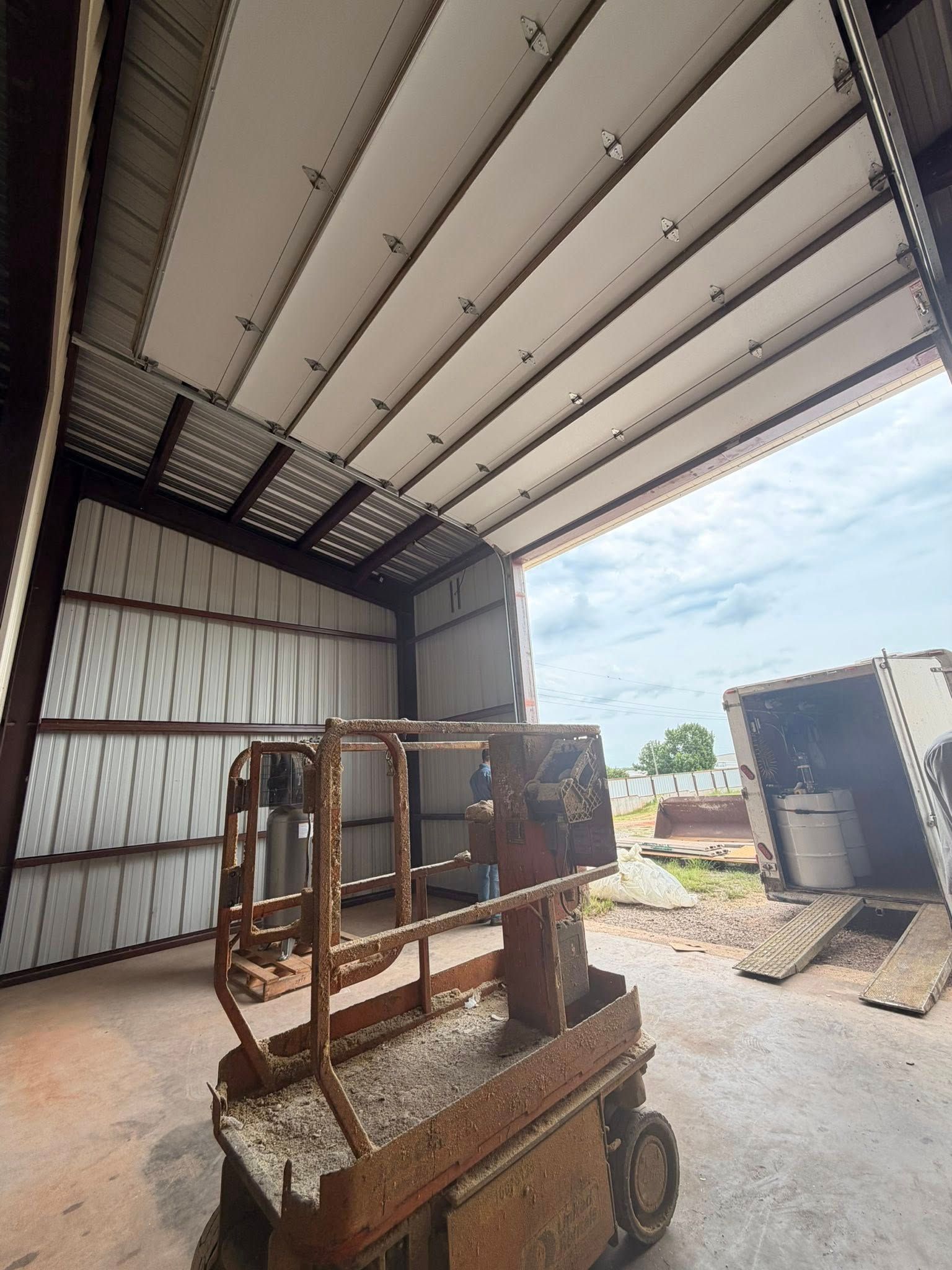 Warehouse interior with a rusty scissor lift under a metal roof, open to daylight outside