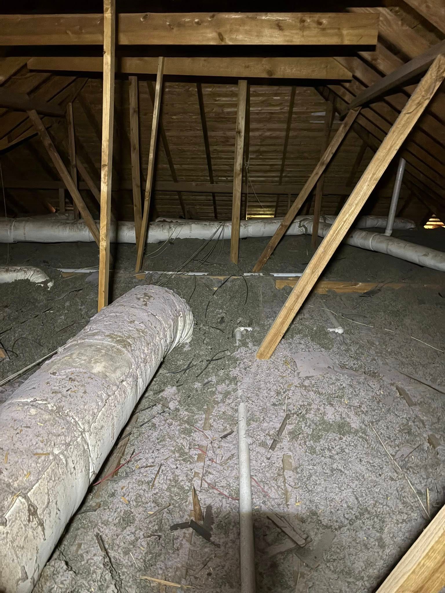 Attic interior with exposed wooden rafters and insulation, lit by a flashlight beam.