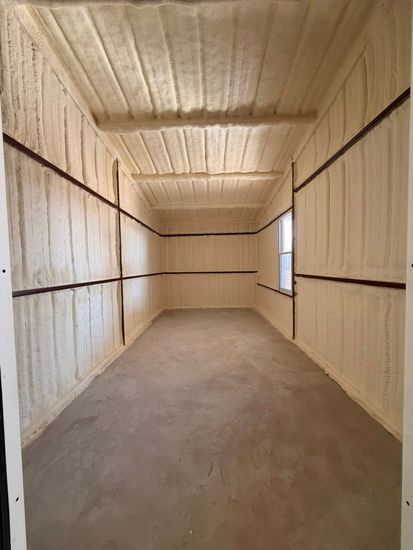 Long workshop interior with wood trusses, a workbench, shelving, and boxes under bright ceiling lights
