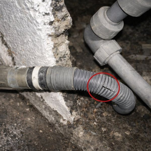 Damaged flexible gray pipe with a crack, near a wall and other plumbing.