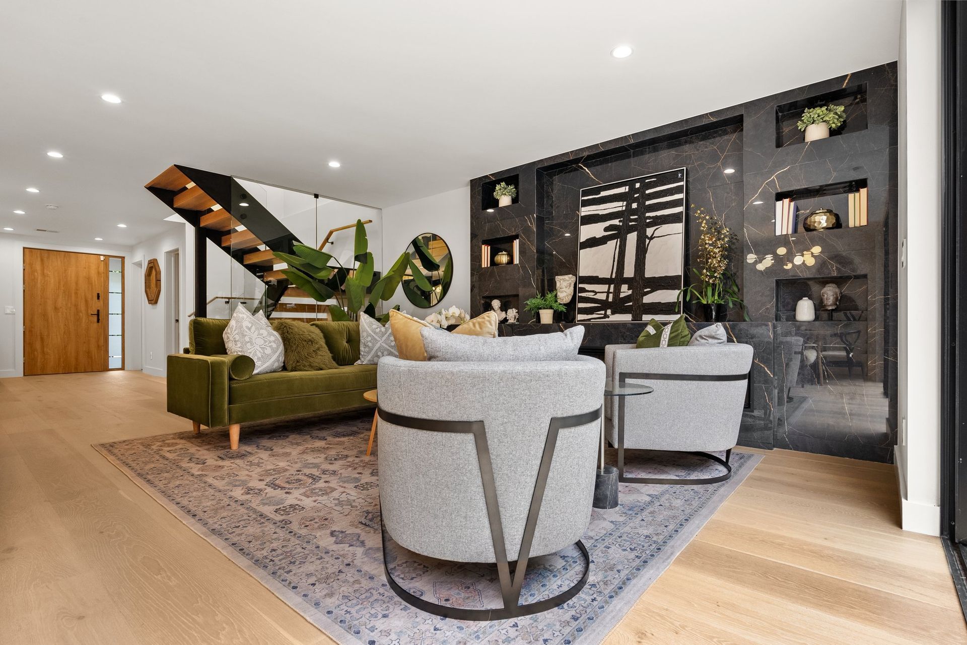 Modern living room with green sofa, white chairs, staircase, and dark built-in shelving.