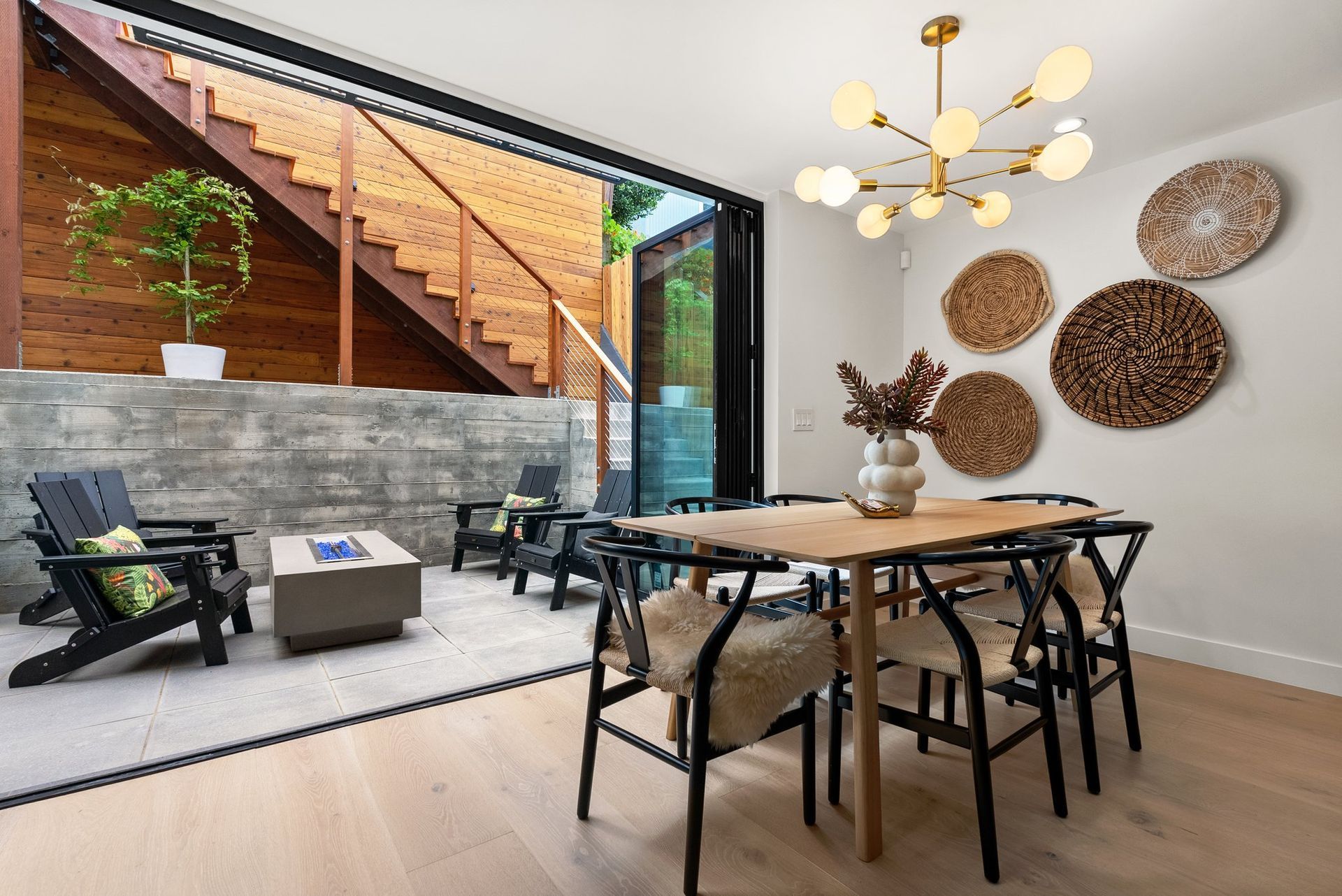 Modern dining area with black chairs, wood table, woven wall decor, and adjacent patio seating under stairs