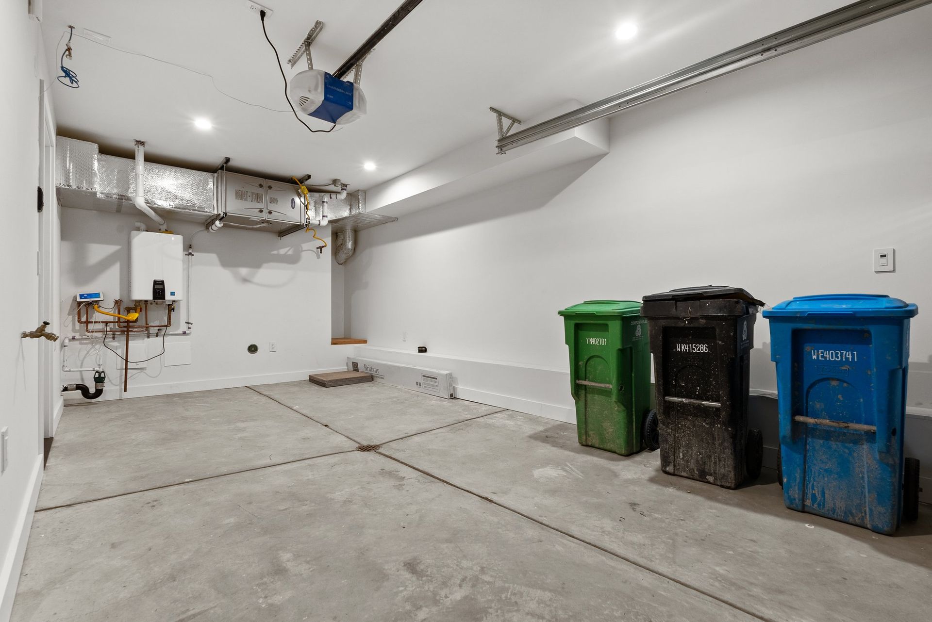 Empty garage with concrete floor, white walls, overhead door track, and three trash bins along the right wall.