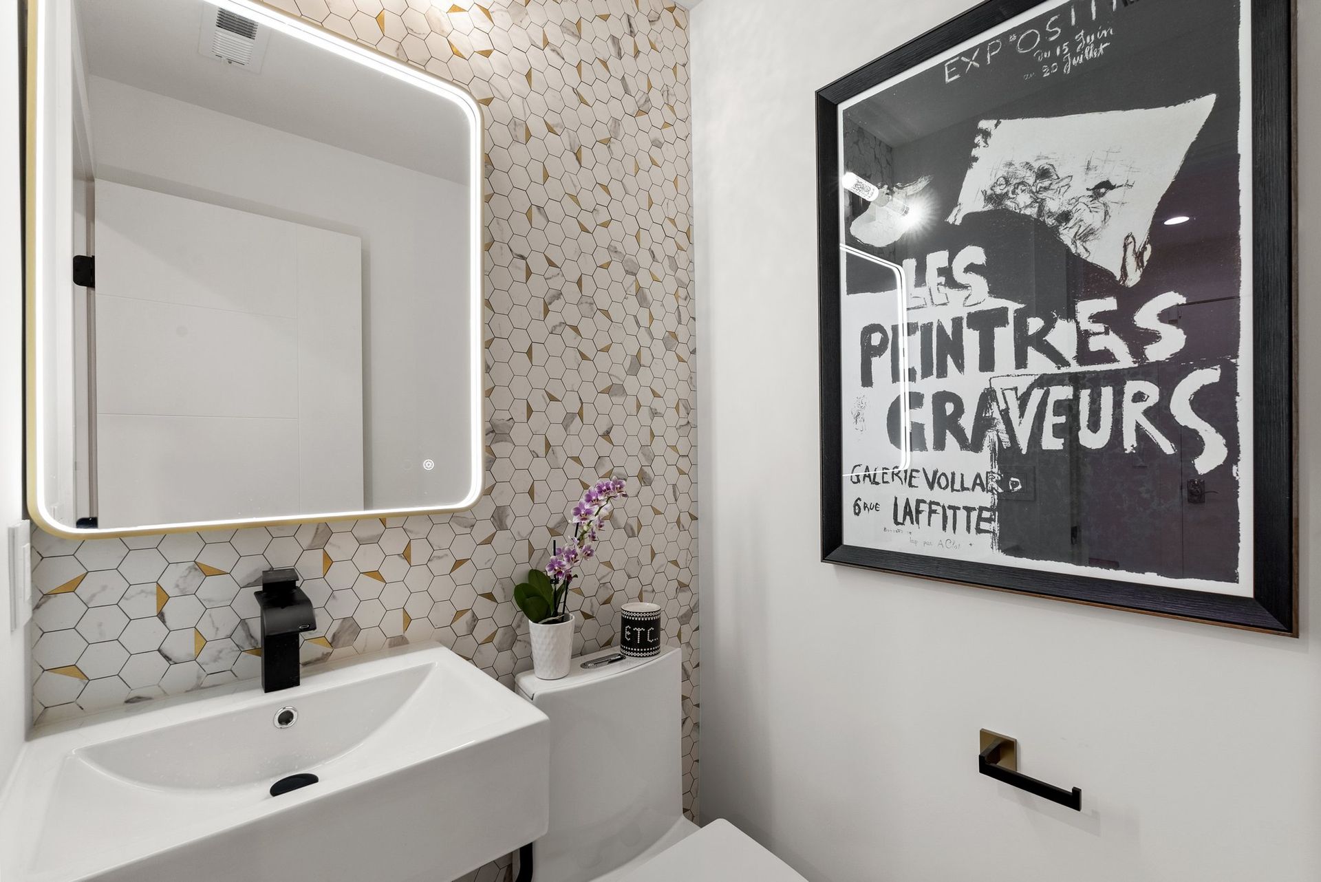 Small modern bathroom with lit mirror, white sink, flowers, and framed black-and-white wall art.