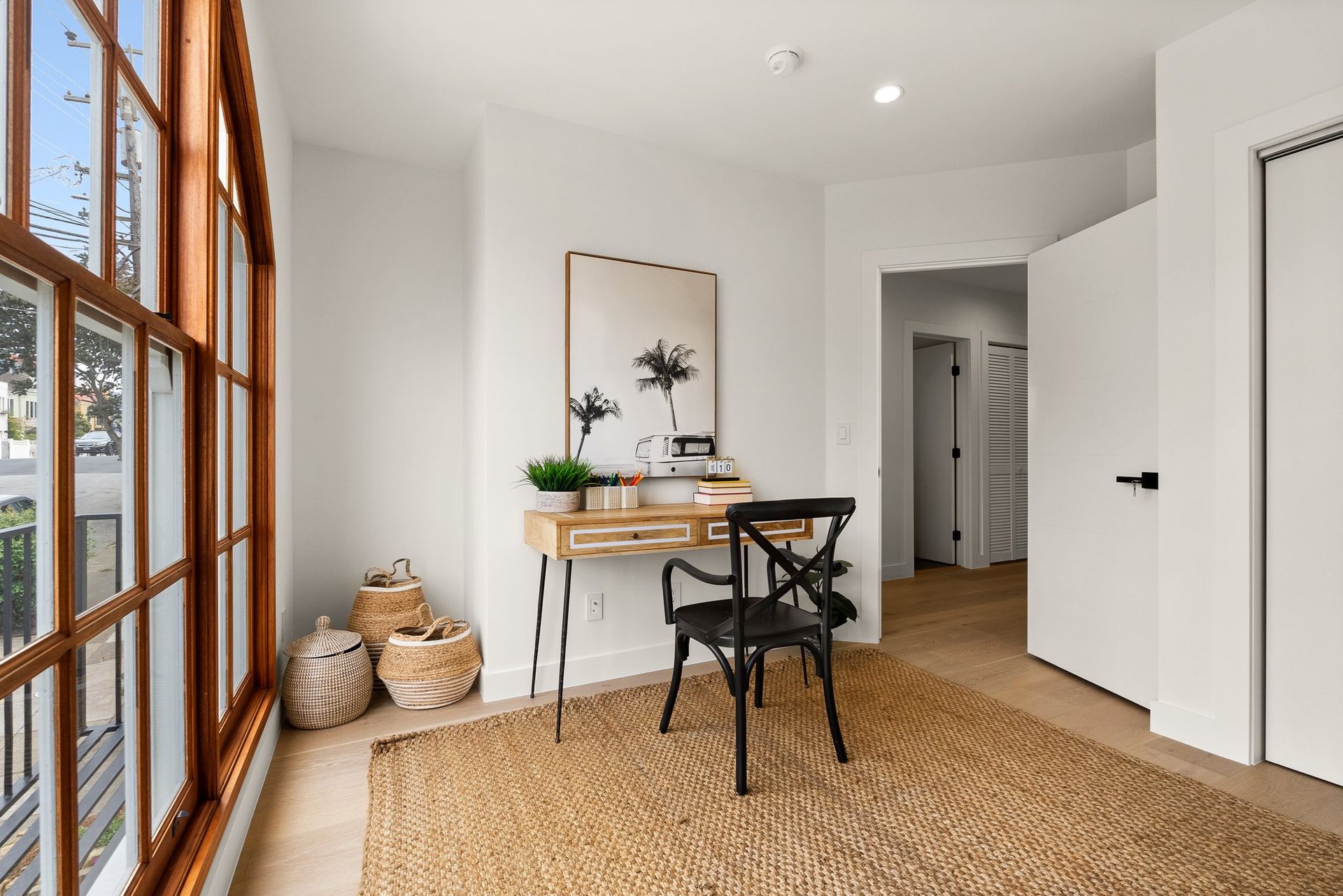 Bright home office with a wooden desk, black chair, woven rug, baskets, and large windows.