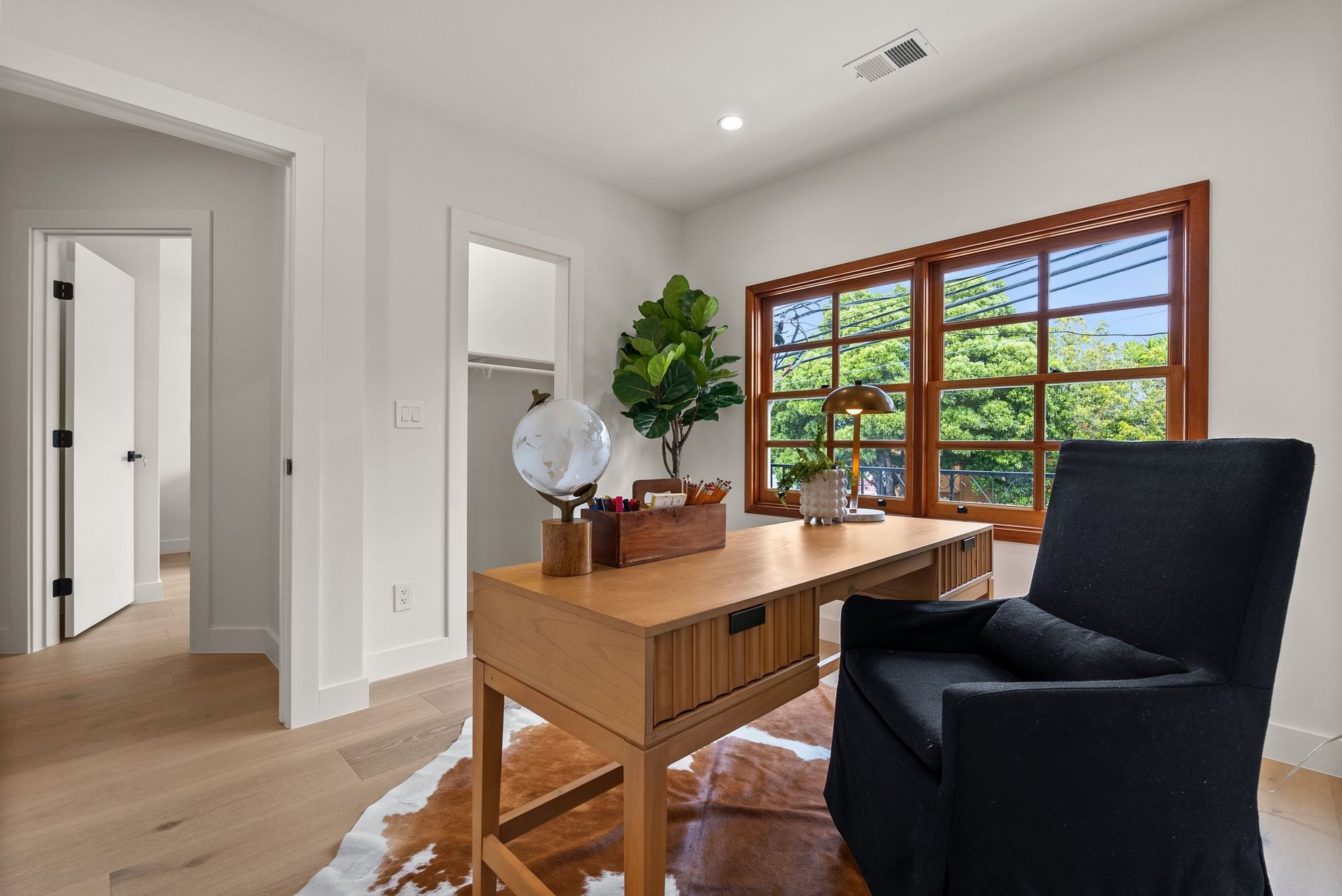 Home office with wooden desk, black chair, large window, and a potted plant in a bright room