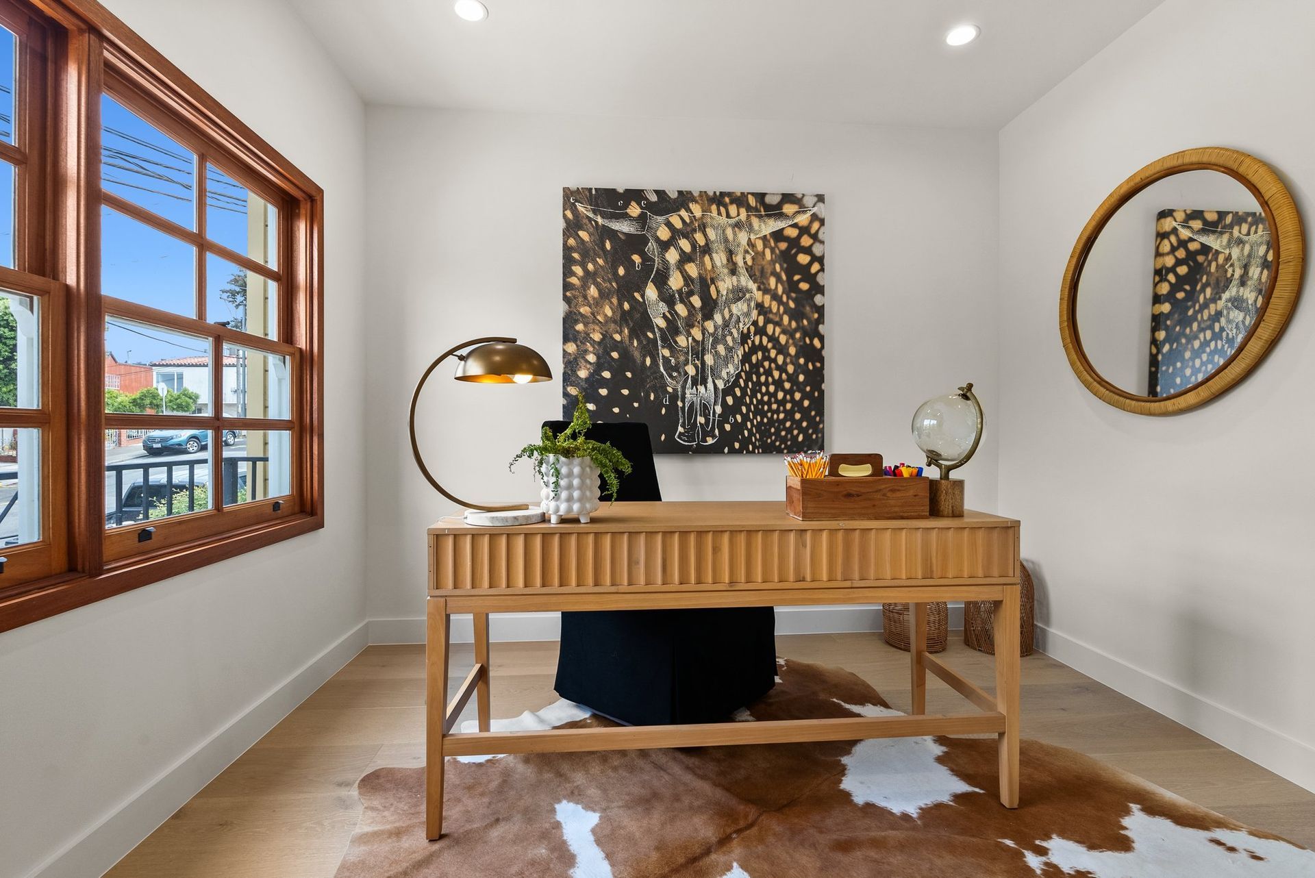 Modern home office with wooden desk, round mirror, lamp, art, and cowhide rug by a window