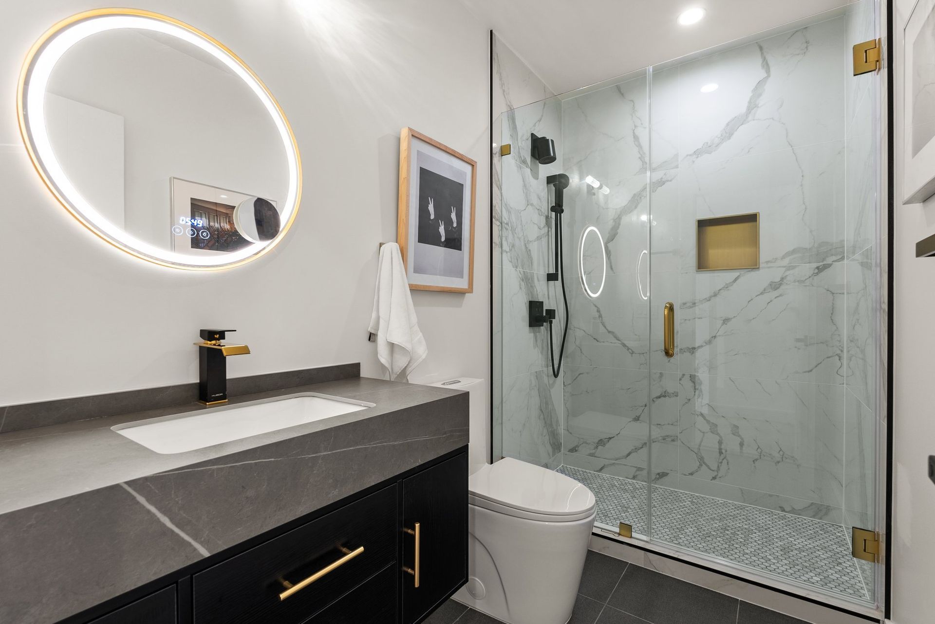 Modern bathroom with vanity, round lit mirror, toilet, and glass walk-in shower.