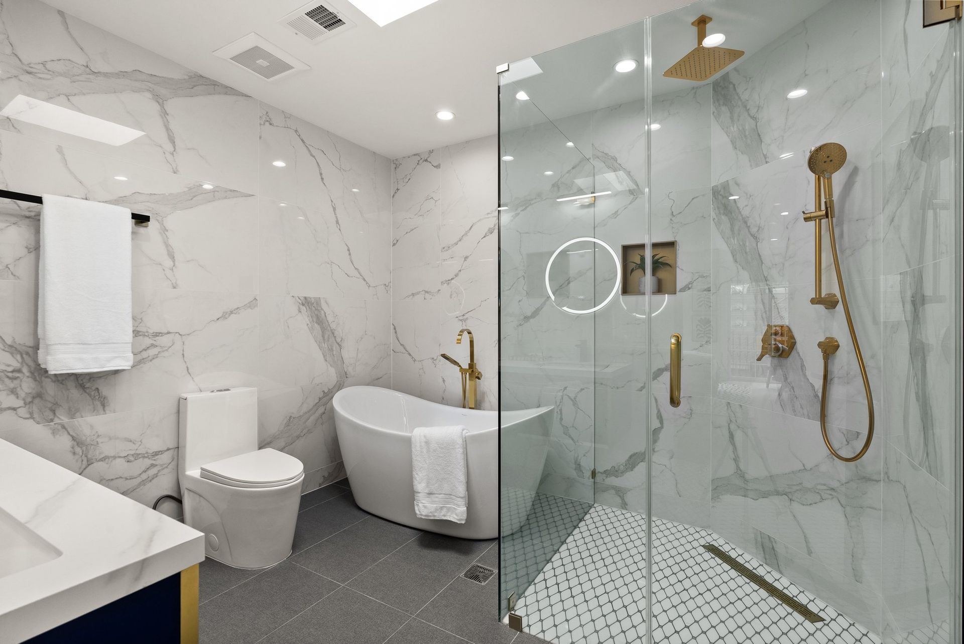 Modern white bathroom with marble walls, glass shower, freestanding tub, toilet, and gold fixtures