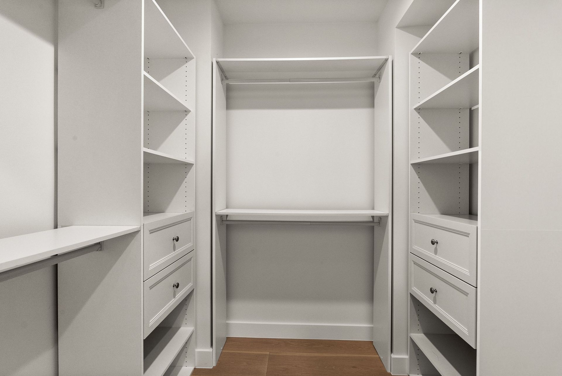 Empty white walk-in closet with built-in shelves, drawers, and hanging rods, and a wood floor.
