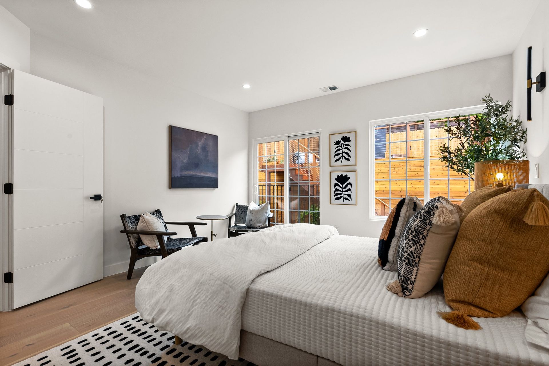 Bright bedroom with a bed, desk, and large windows, decorated in neutral tones and warm light