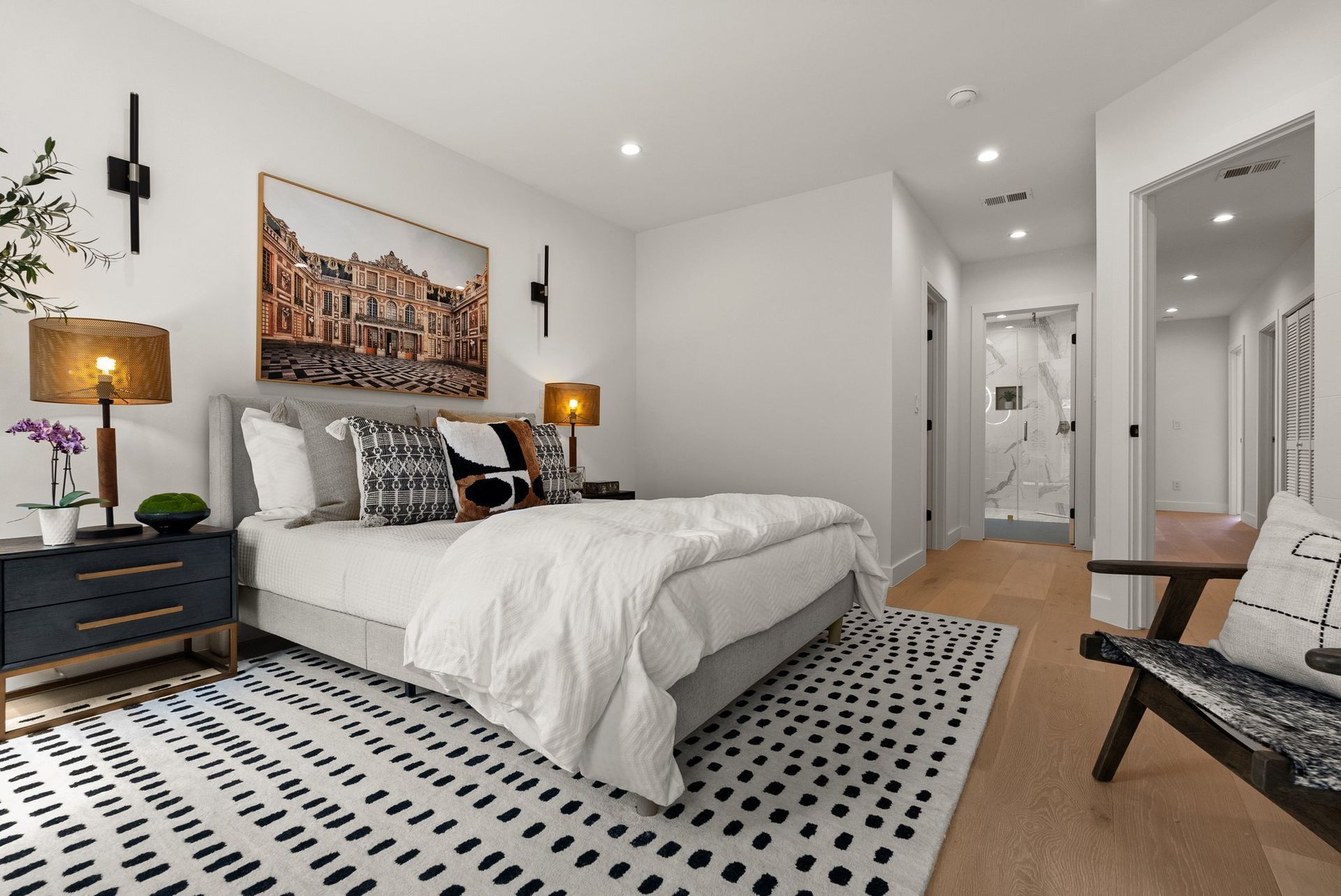 Modern bedroom with white bedding, patterned rug, bedside lamps, wall art, and a hallway view.