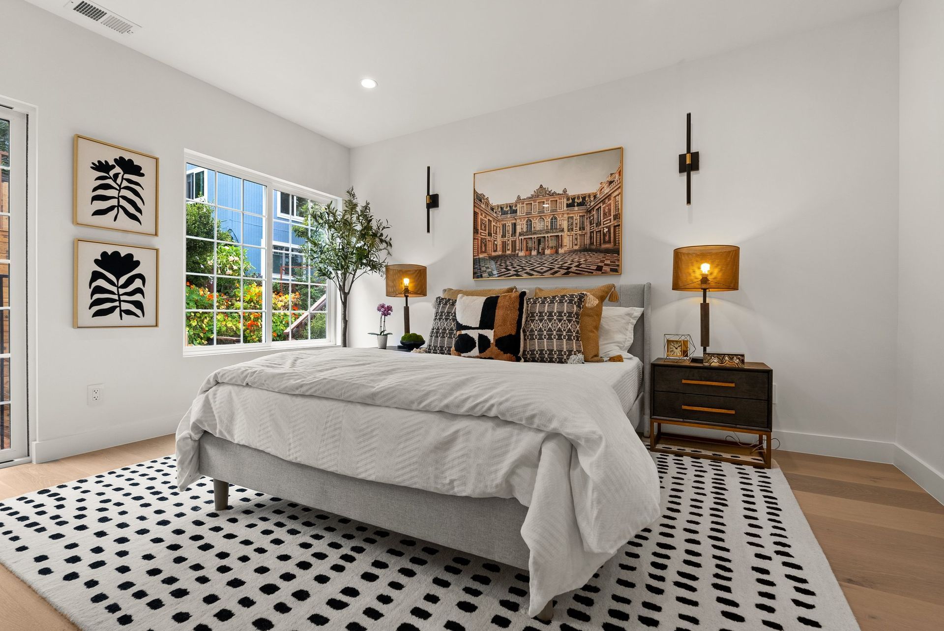 Bright modern bedroom with a bed, patterned rug, nightstands, lamps, and wall art.