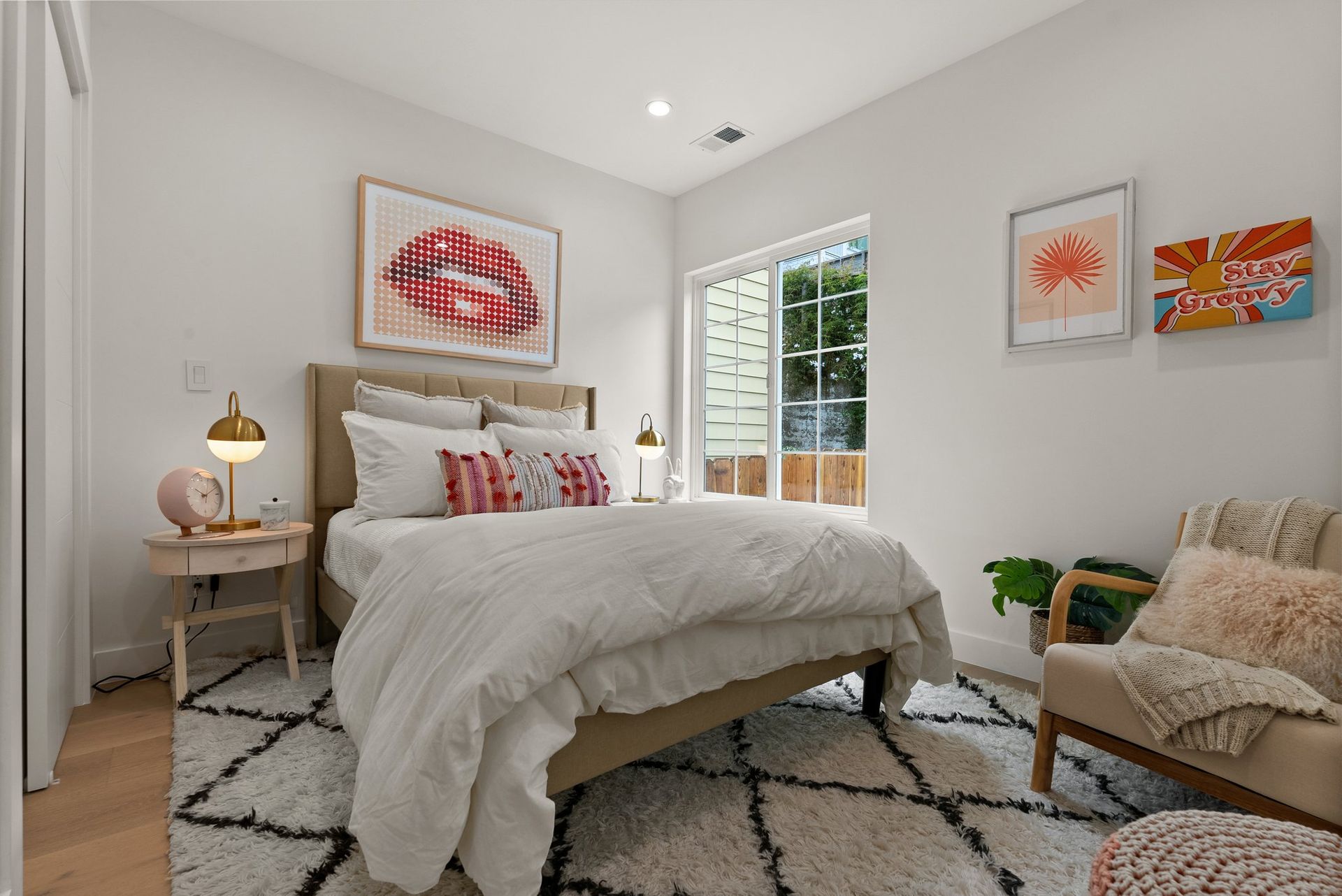 Bright bedroom with a bed, patterned rug, bedside lamp, and framed wall art near a window.