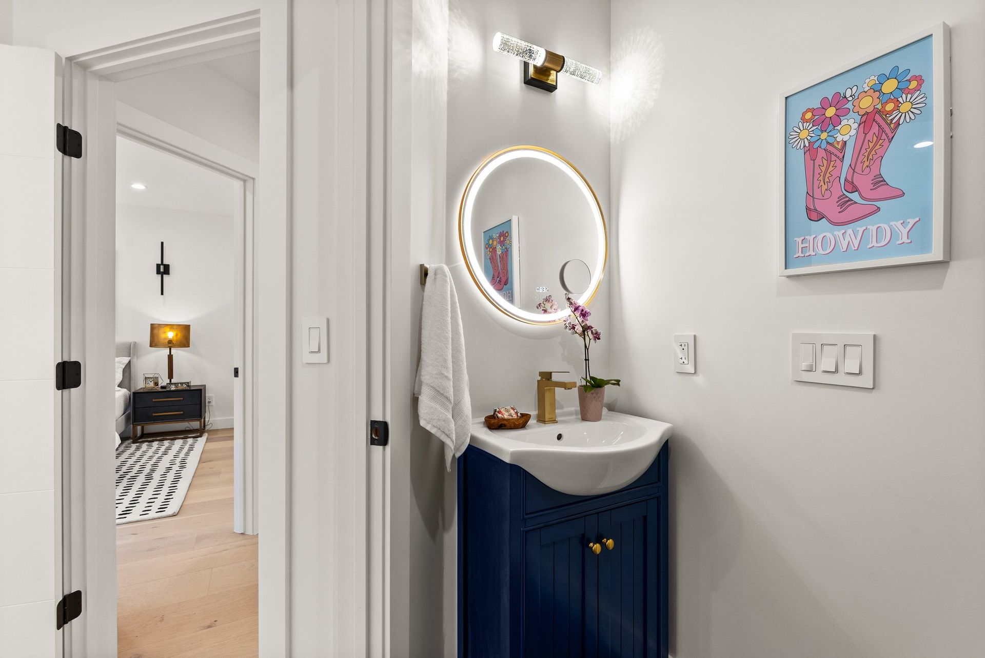 Small hallway vanity with round lit mirror, blue cabinet, and colorful wall art.