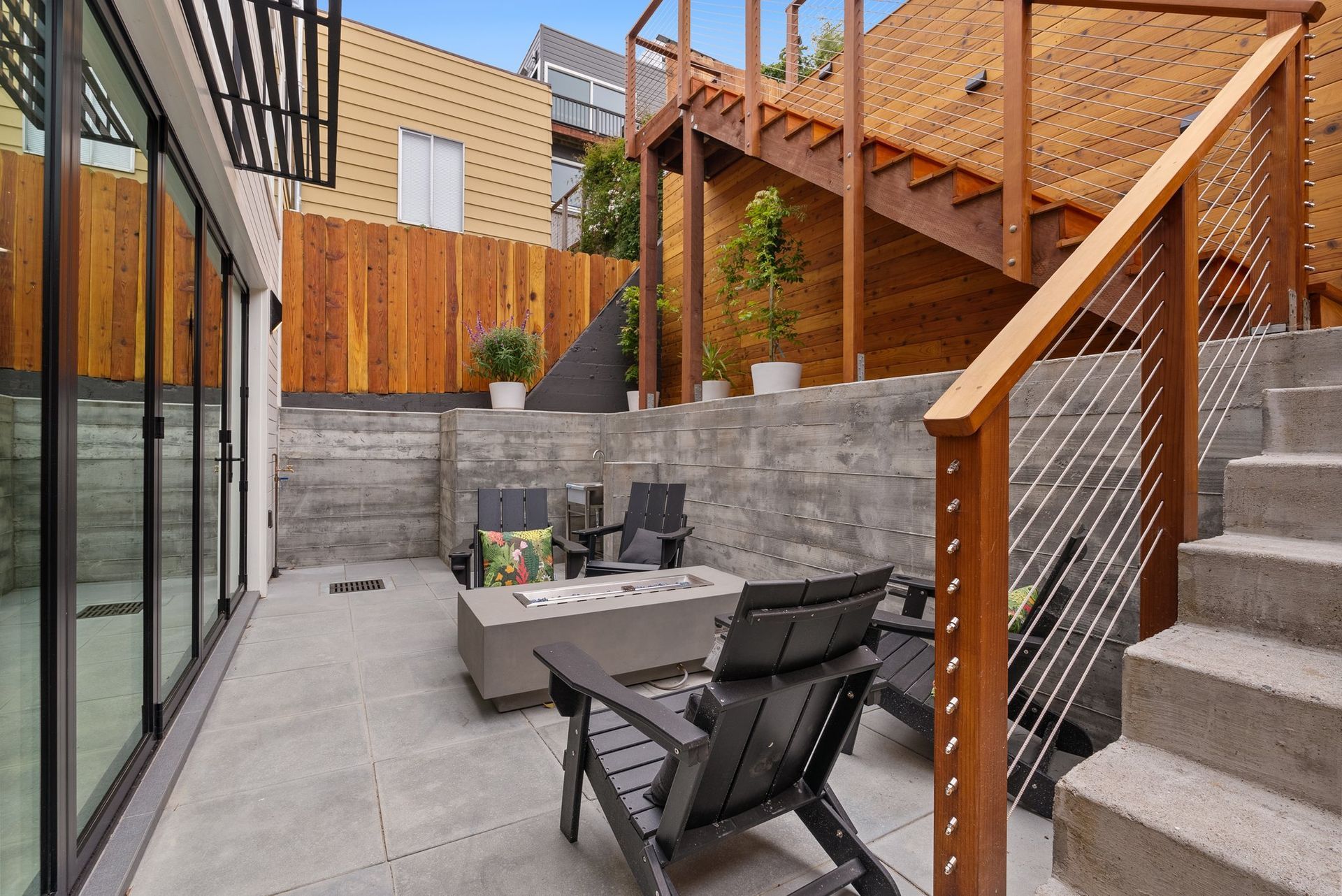 Modern backyard patio with gray seating, fire pit, wood fence, and stairs beside a glass house