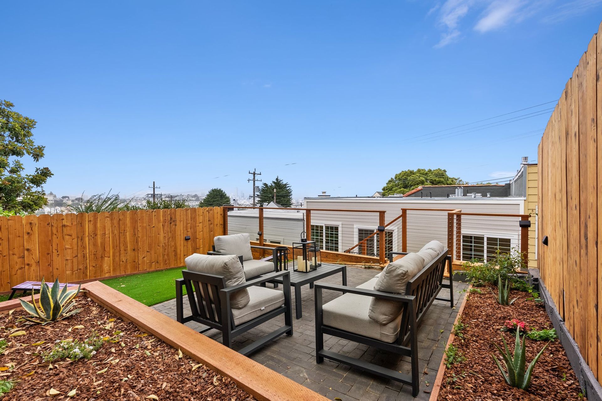 Patio seating area with gray sofa, chairs, fire pit, and wooden privacy fences under a blue sky