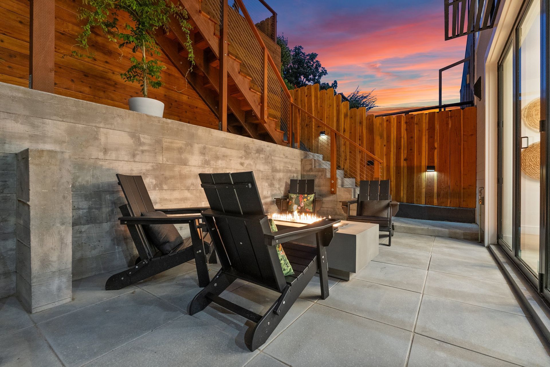 Modern backyard patio with two black lounge chairs, fire pit, wood fencing, and sunset sky