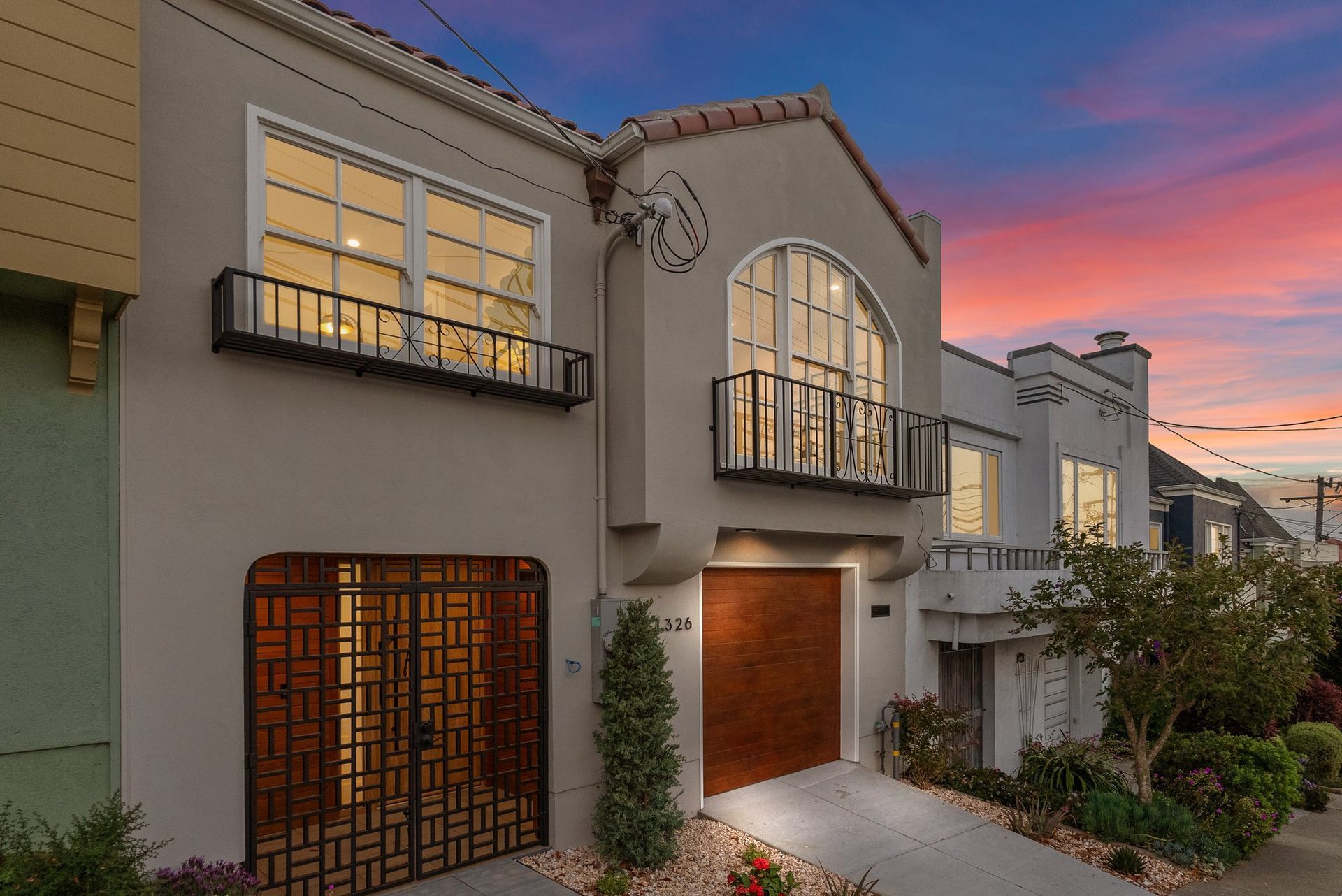 Modern townhouse at dusk with lit windows, arched balconies, and a sunset sky over a landscaped entryway