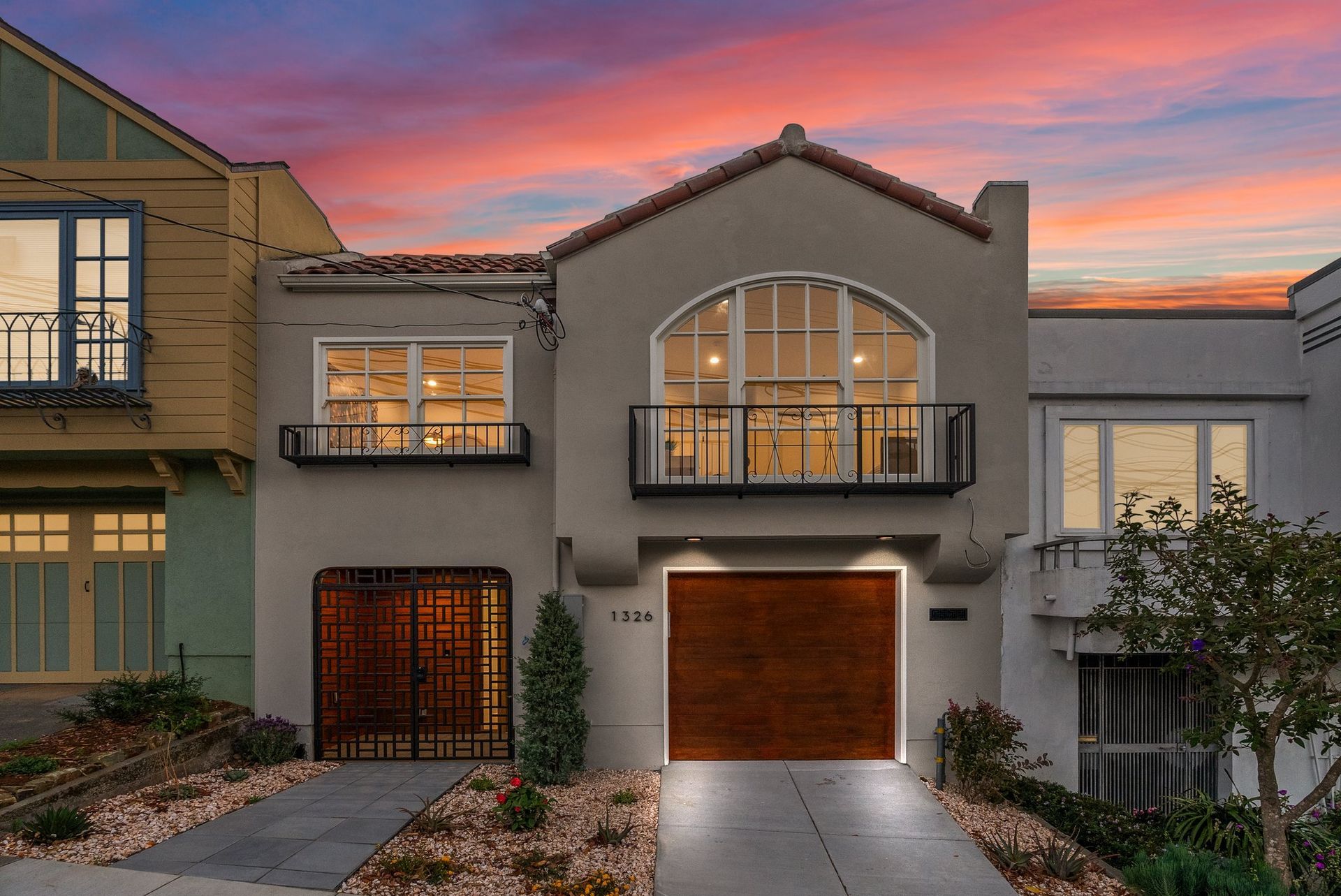 Modern townhouse exterior at sunset with lit windows, balcony, and garage doors