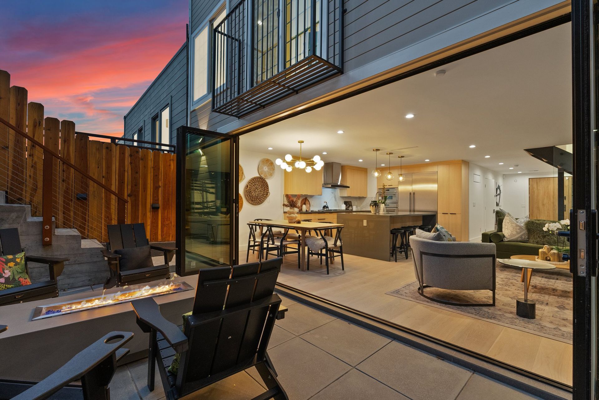 Modern patio opening to a lit kitchen and living room at sunset, with outdoor dining and lounge seating