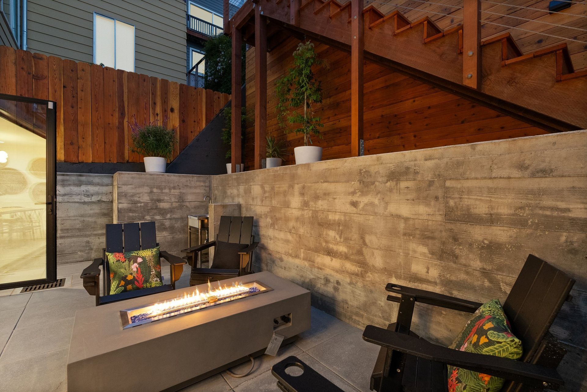 Modern patio with concrete fire table, black chairs, stone wall, and warm wood fencing at dusk