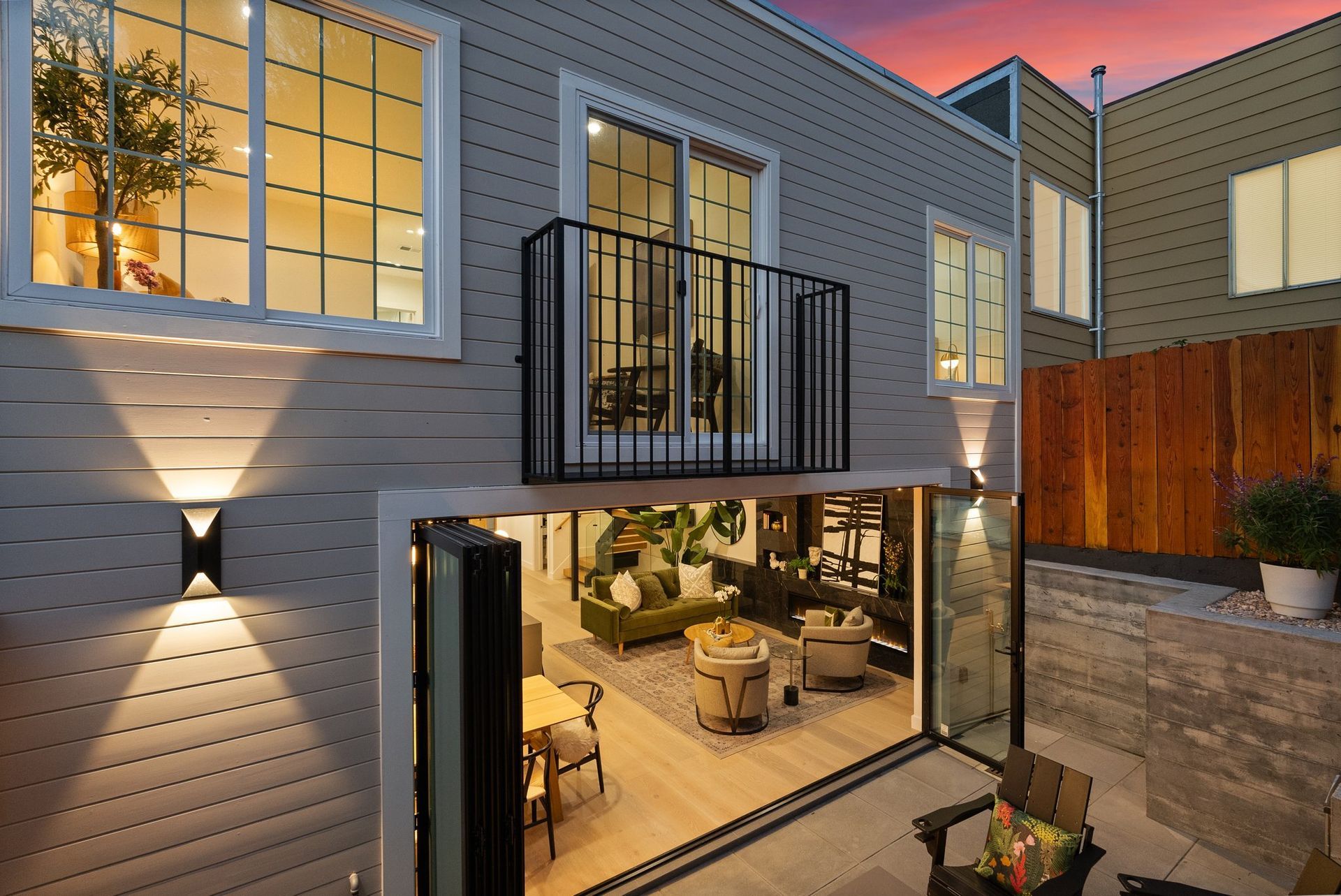 Modern house exterior at dusk with lit basement patio and upstairs balcony, framed by brick and wood fencing