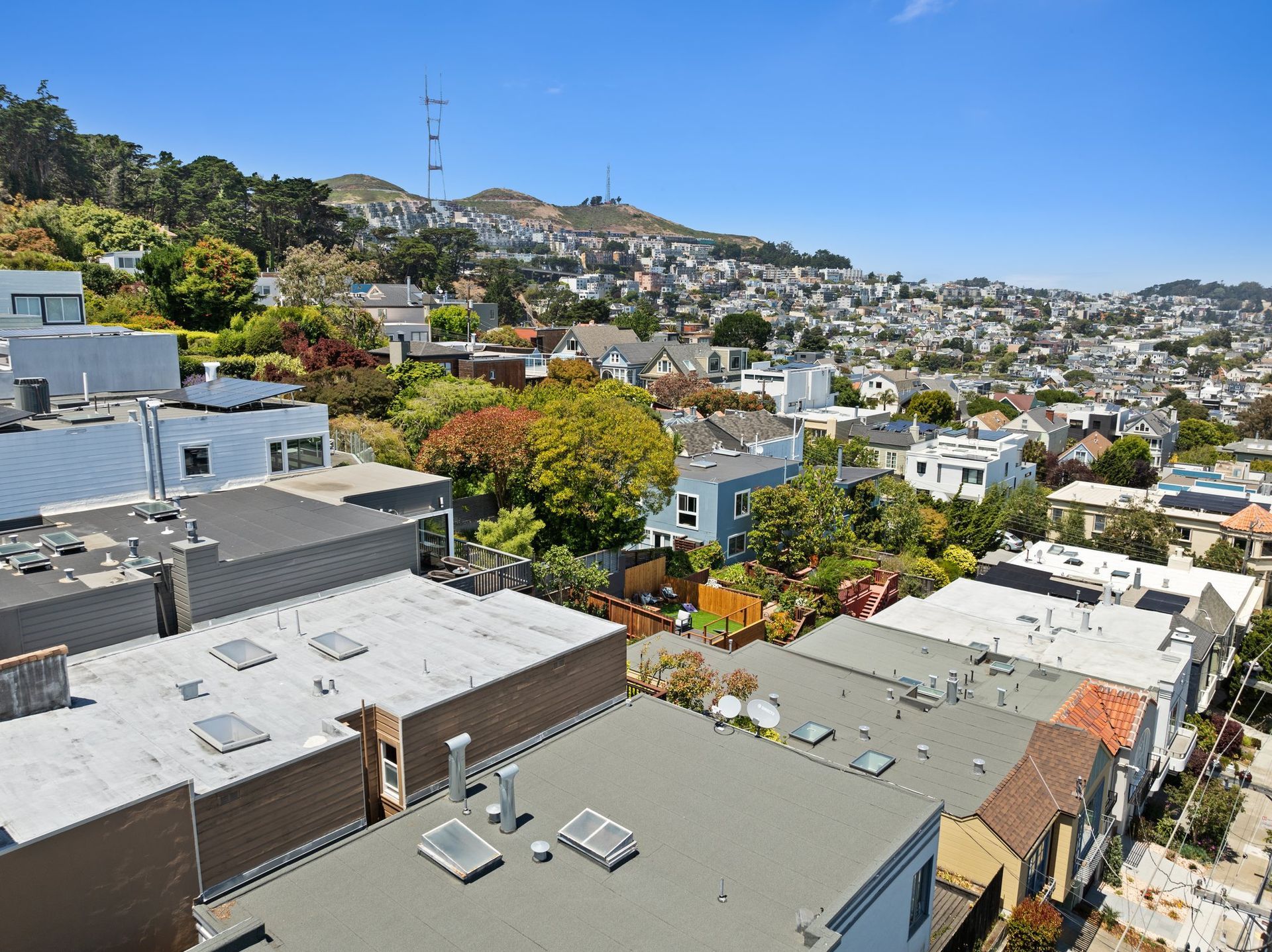Hillside neighborhood of white houses under a clear blue sky, with rooftops and trees in the foreground
