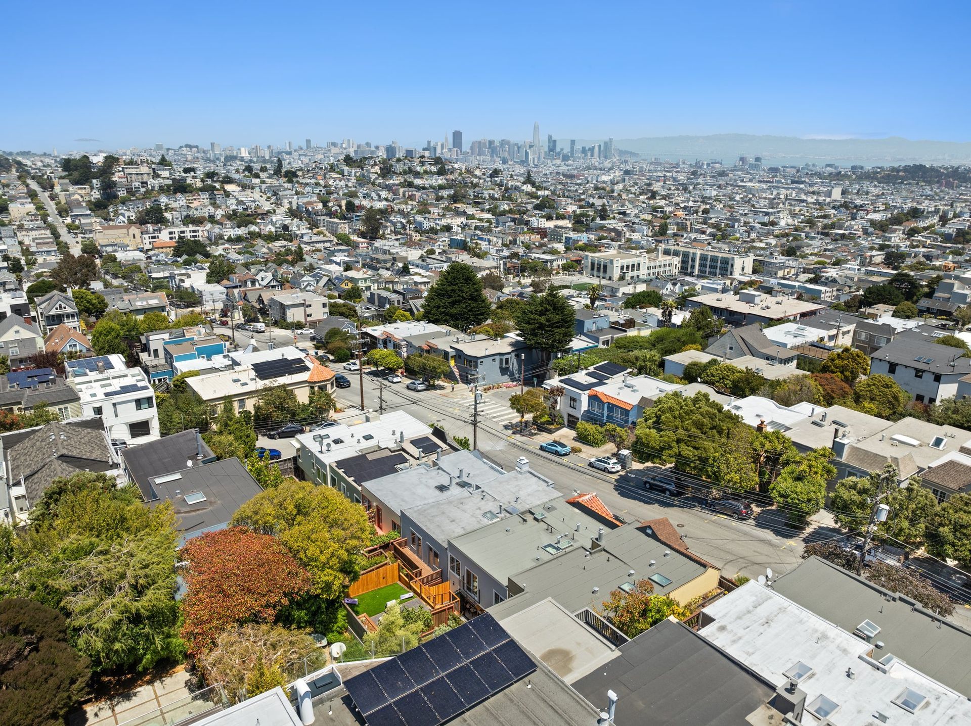 Aerial view of a dense city neighborhood with rooftops, trees, and distant skyline under a clear sky