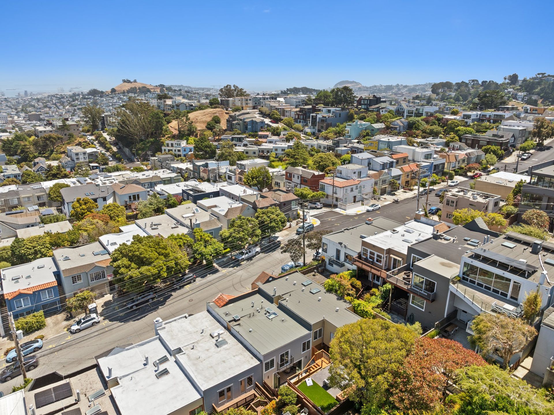 Aerial view of a hilly neighborhood with white houses, trees, and a blue sky.