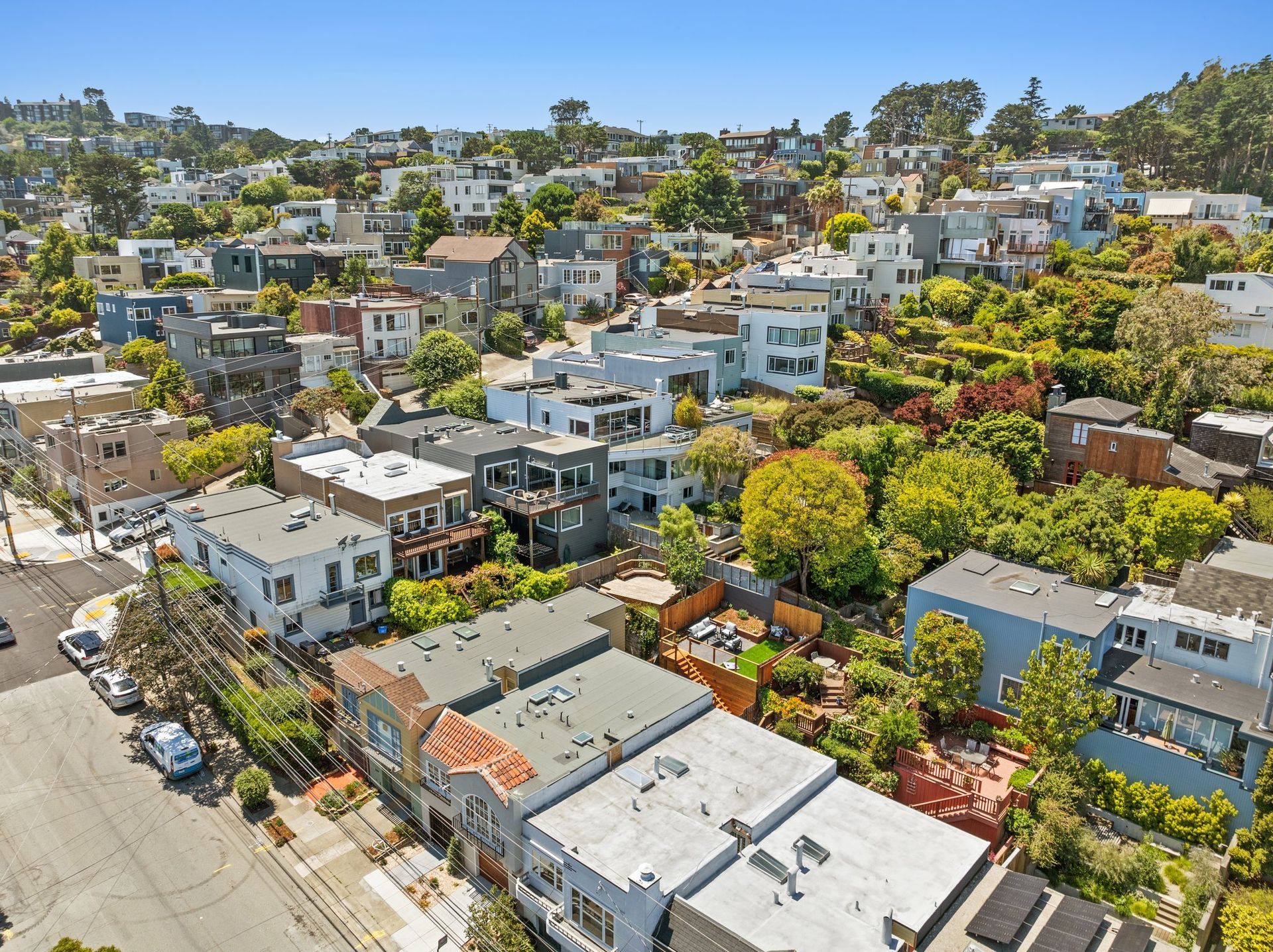 Aerial view of a dense hillside neighborhood with white houses, trees, and a clear blue sky.