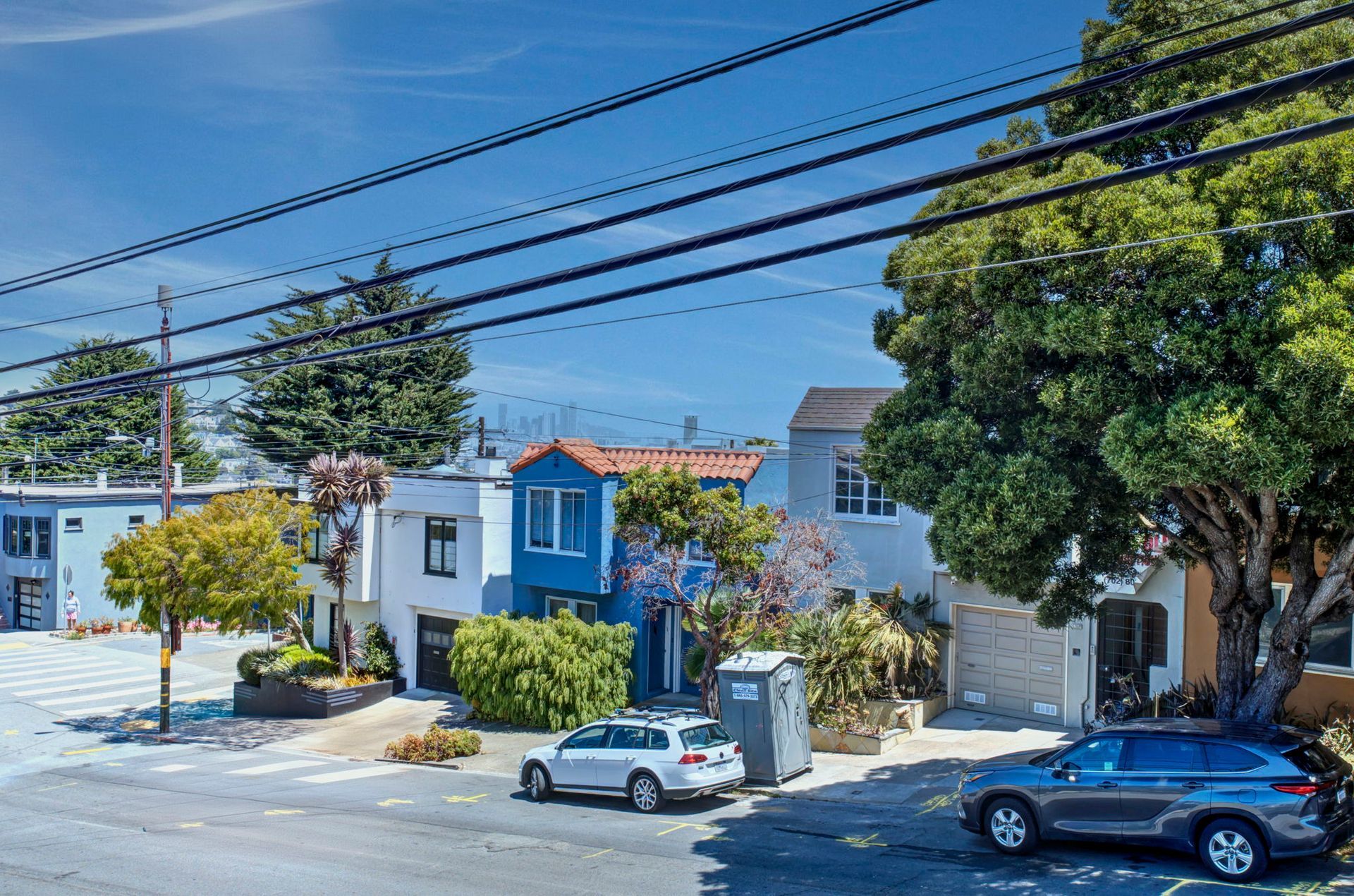 Quiet residential street with colorful houses, parked cars, power lines, and a large tree in bright sun