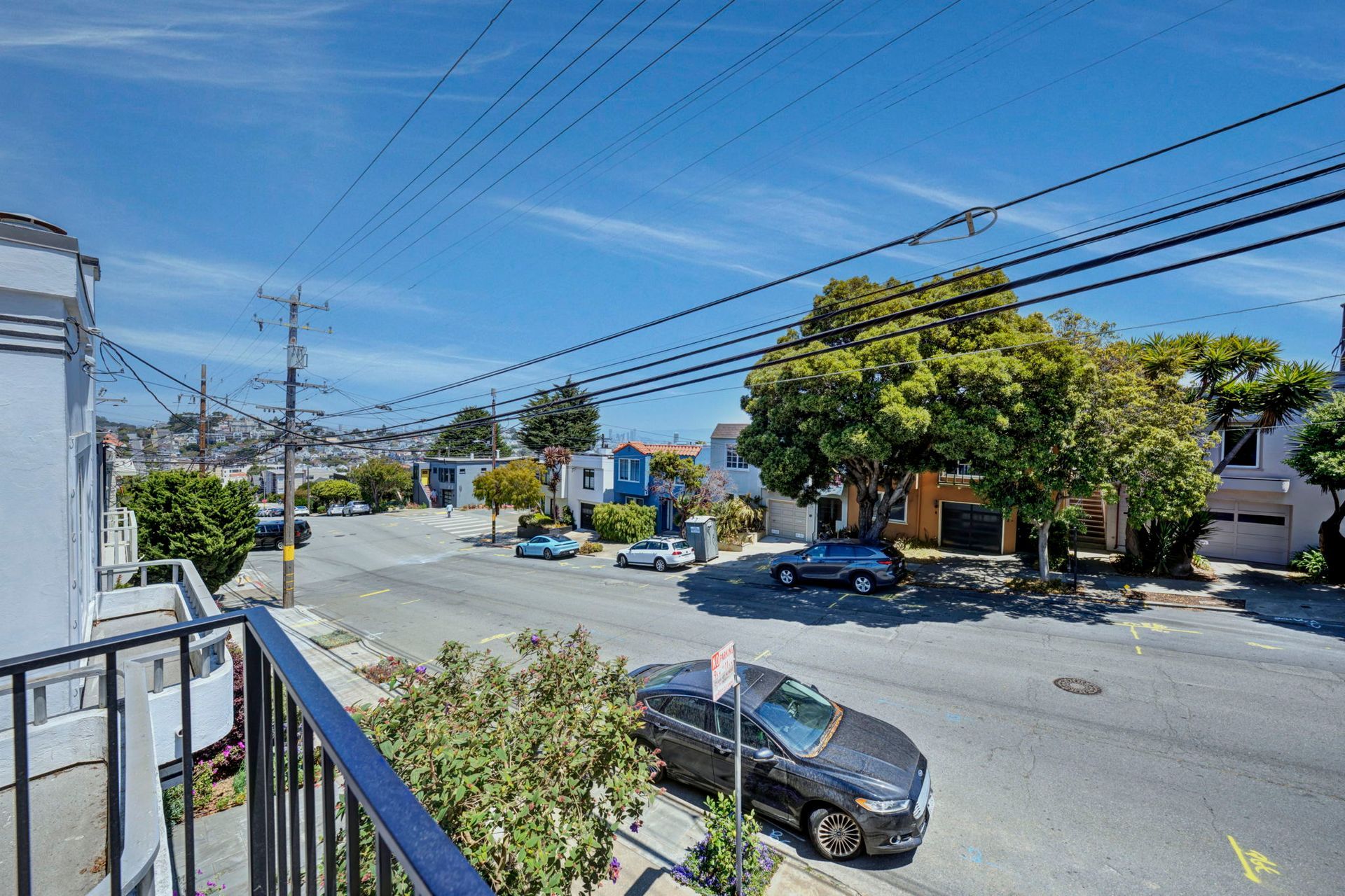 Sunny street view from a balcony, with parked cars, trees, power lines, and houses under a blue sky.
