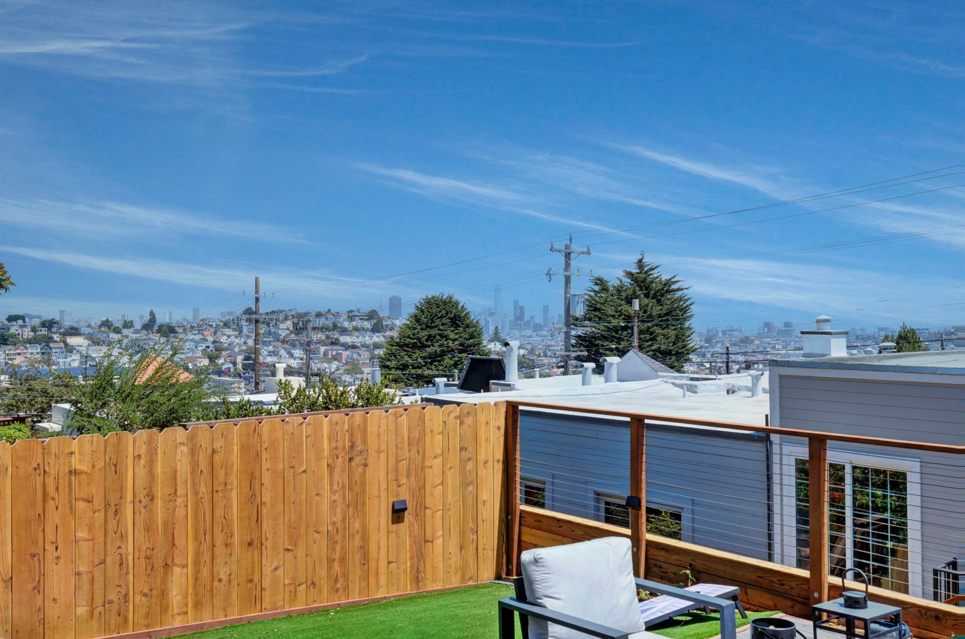 Backyard deck with wooden fence, white chairs, and city skyline under a blue sky