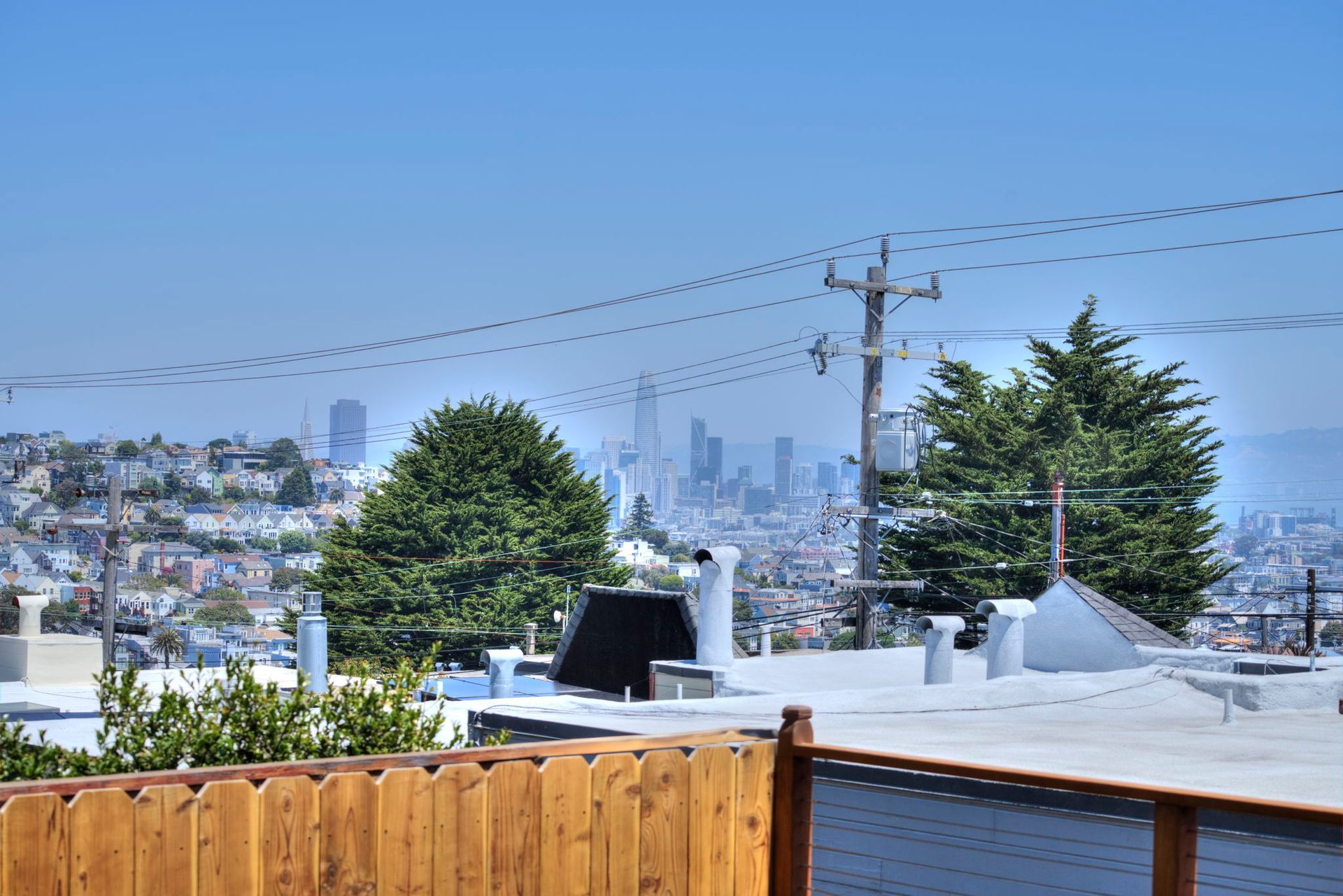 City skyline beyond a wooden fence, with rooftops, trees, and power lines under a clear blue sky