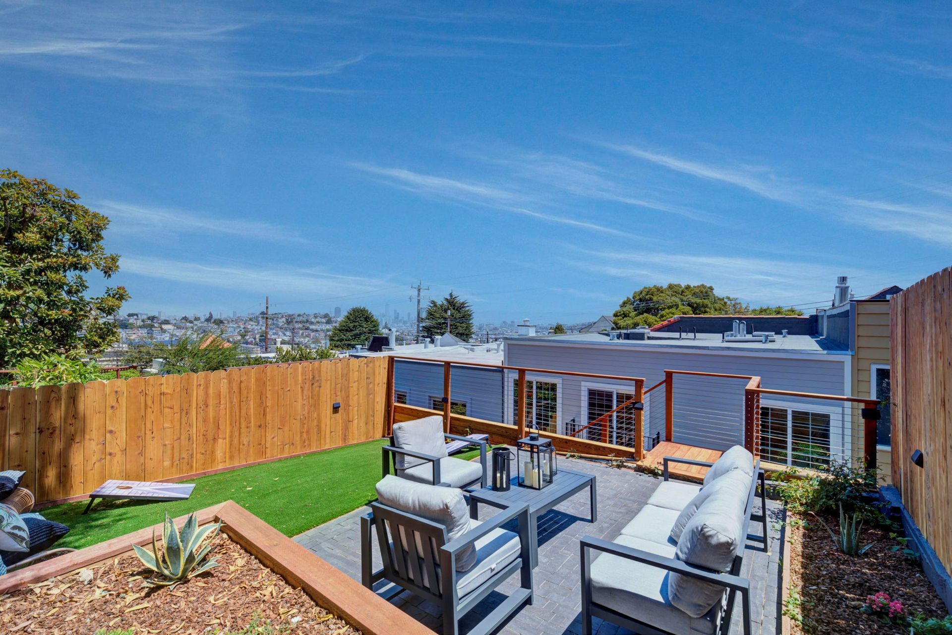 Backyard patio with gray outdoor seating, wooden fence, green lawn, and blue sky view